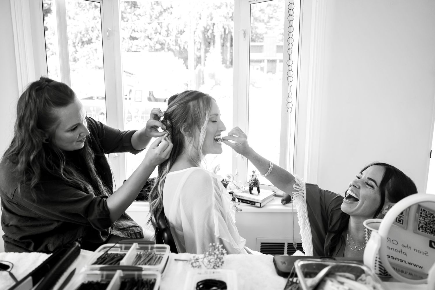 Black and white candid image of people getting ready with hair and makeup near a bright window with laughter and joy.