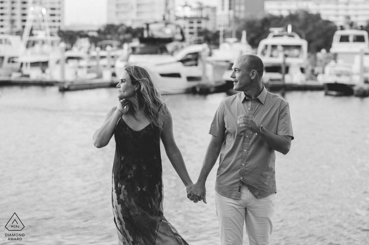 A couple walks hand in hand along a marina waterfront in a romantic black and white engagement photo.