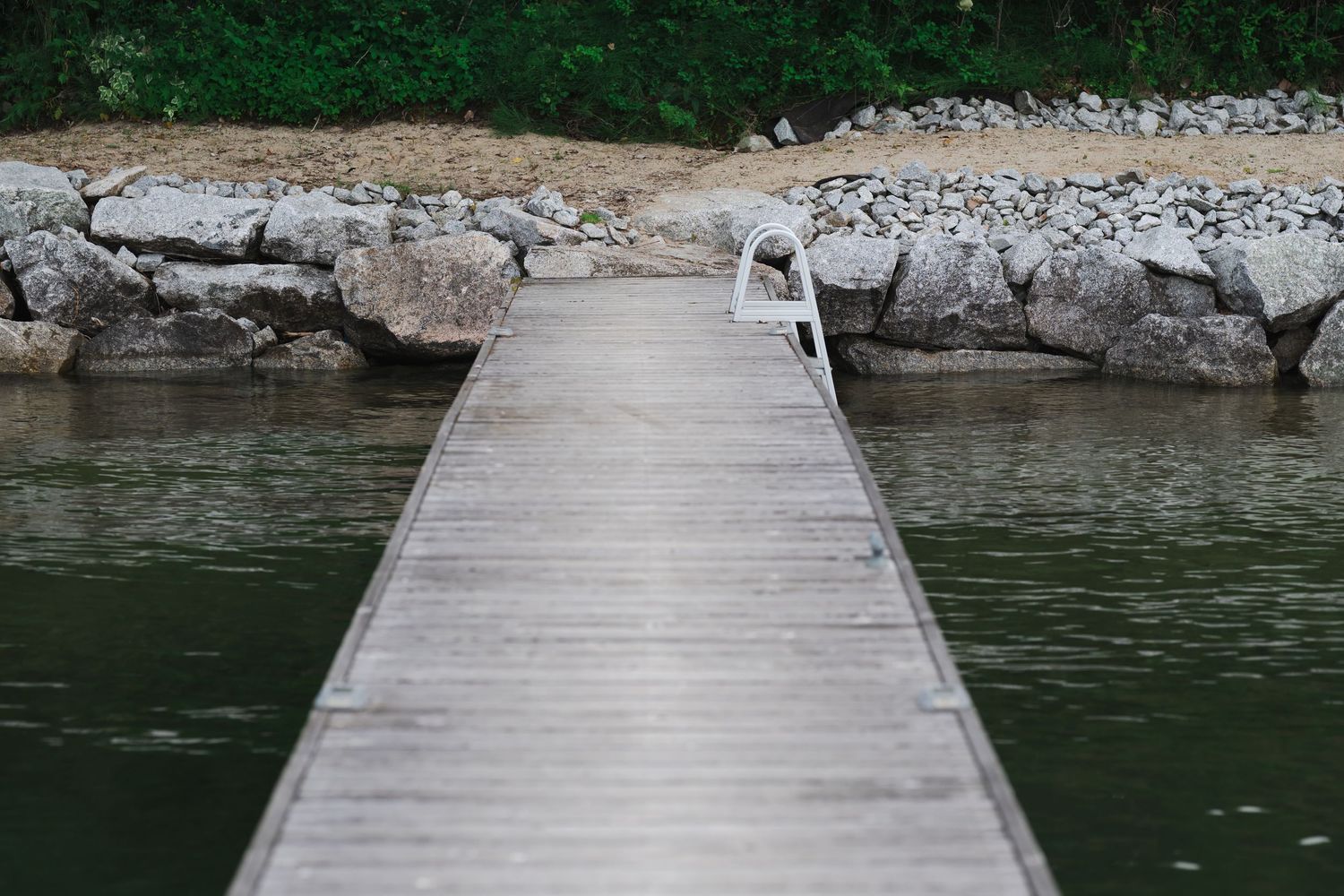 Wooden dock extending over calm green lake water toward rocky shoreline with lush trees in background.