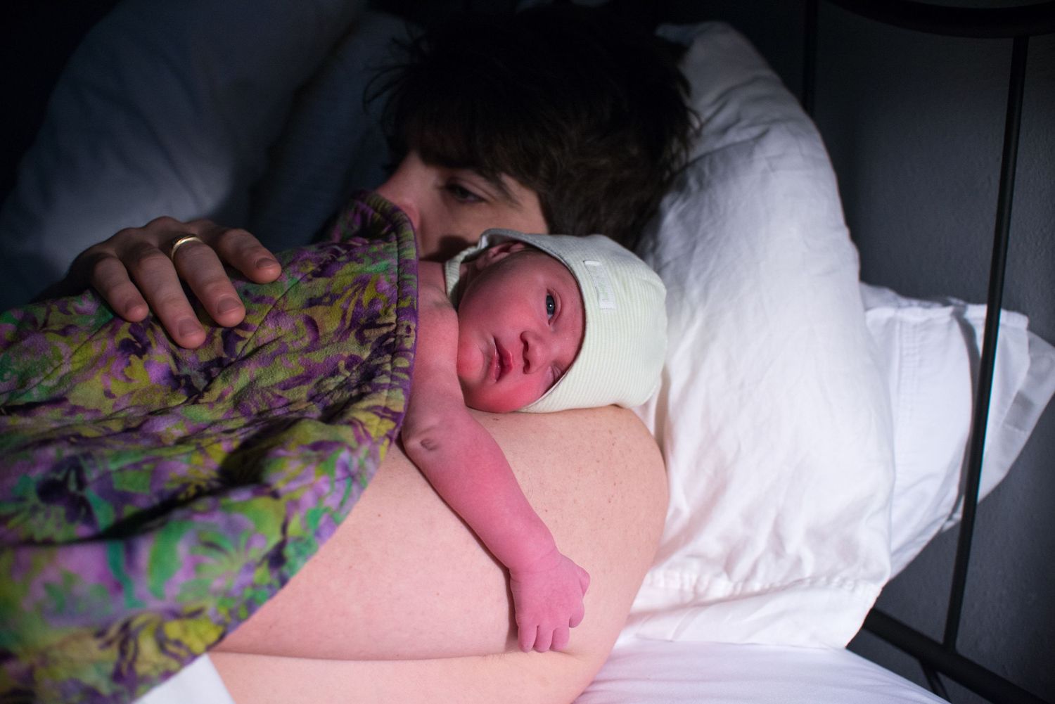 A newborn baby wrapped in a colorful floral blanket rests peacefully on white bedding.