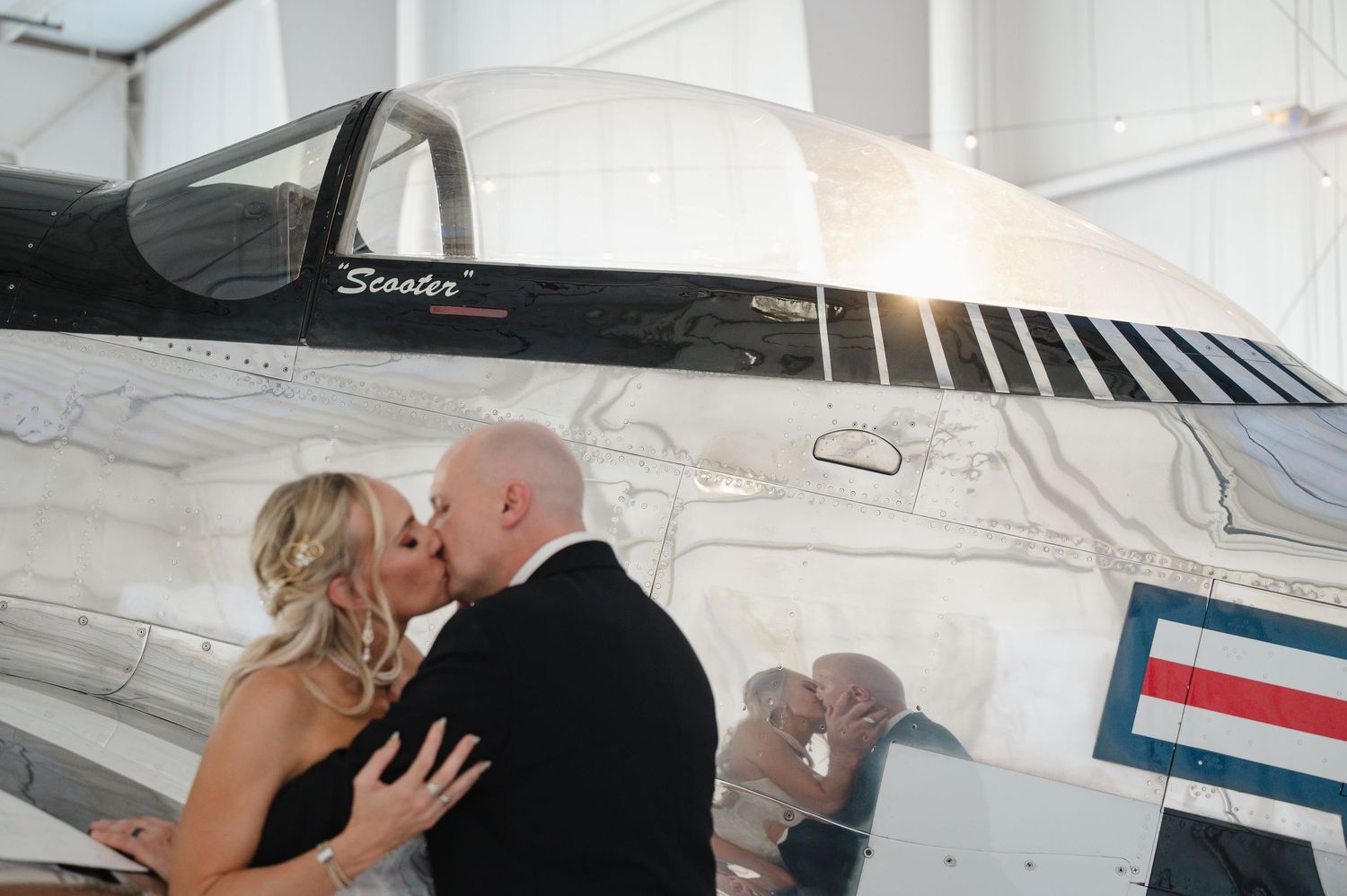 Bride and groom share a passionate kiss beside a vintage military jet in an airplane hangar.