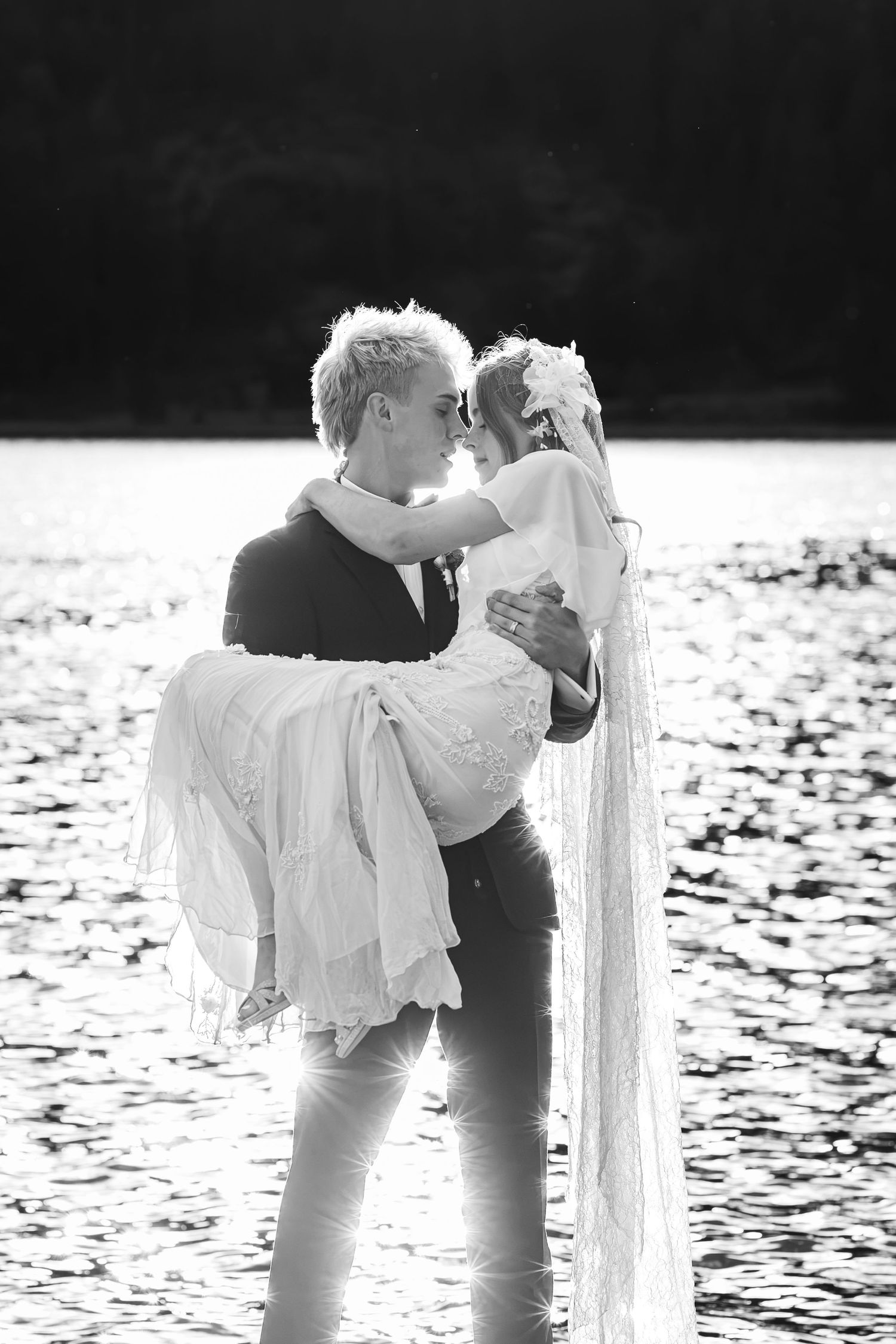 Black and white photo of groom carrying bride near shimmering lake, sharing romantic moment in wedding attire.