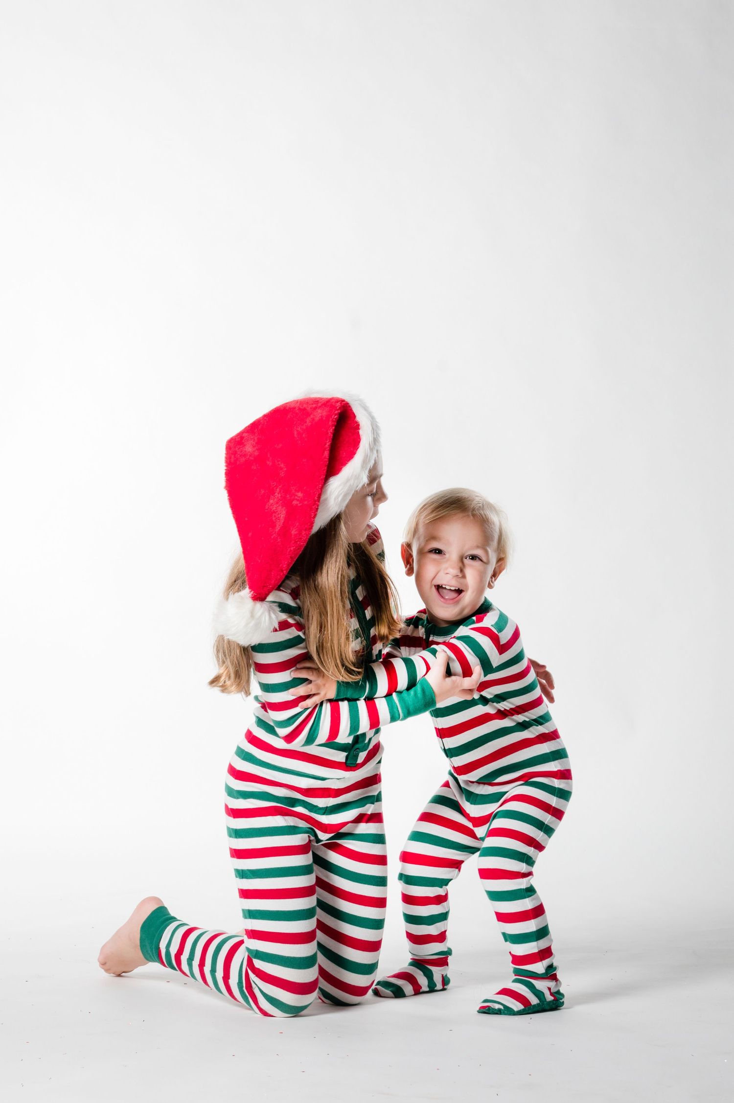 Two children in matching red and green striped Christmas pajamas, one wearing a Santa hat, hugging and laughing together.