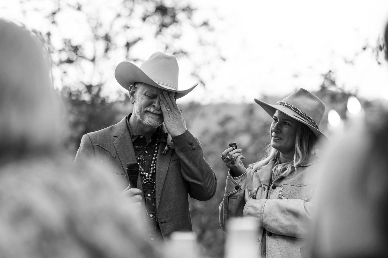 Black and white photo of emotional man in cowboy hat wiping tear at outdoor wedding ceremony.