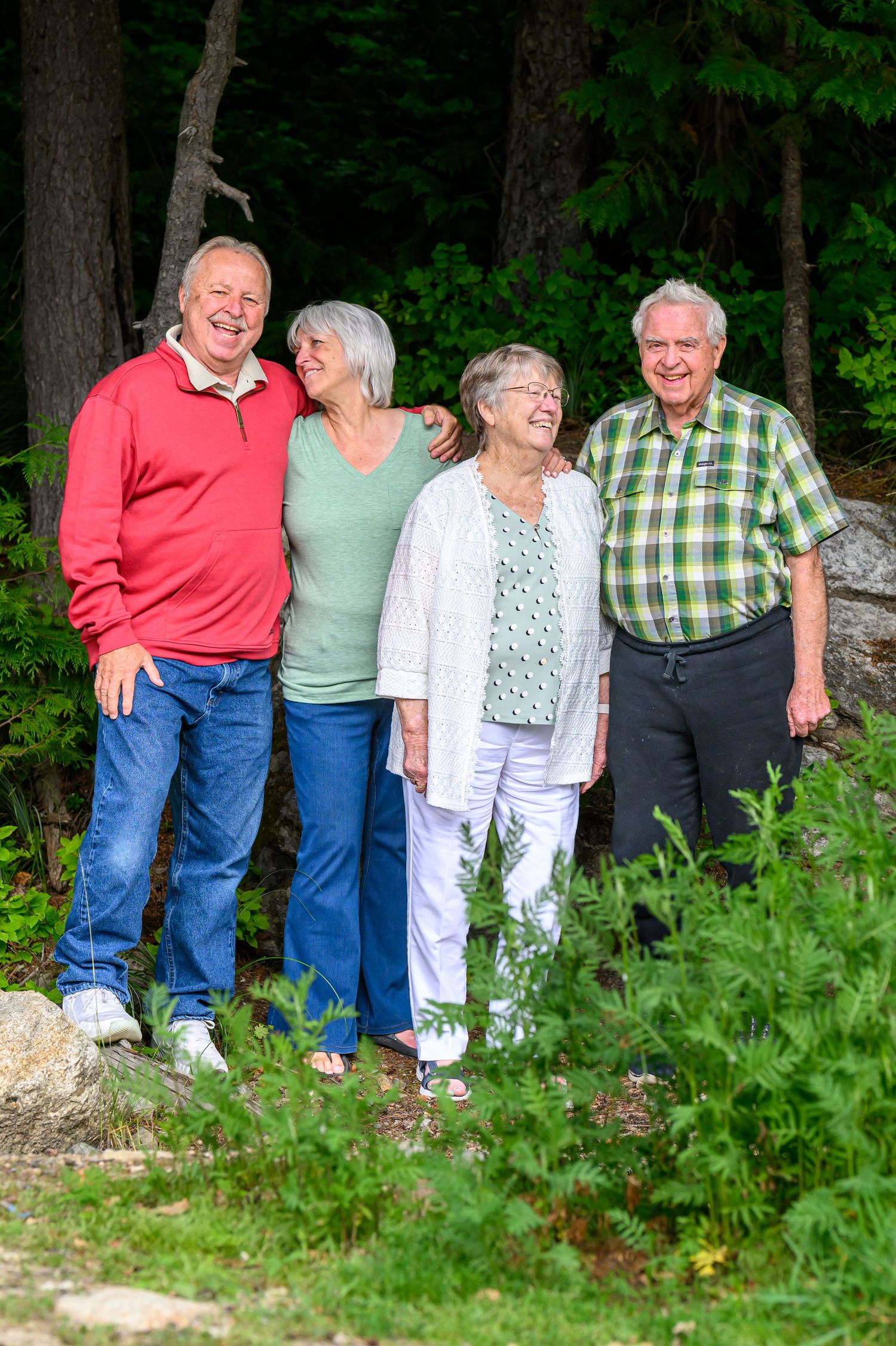 Four cheerful seniors laugh and embrace outdoors surrounded by lush green forest trees.