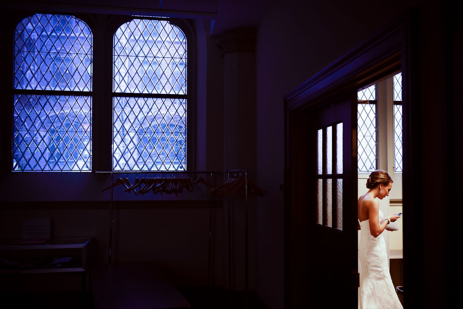 A bride stands in an elegant wedding dress near a stained glass window in a dimly lit church interior.