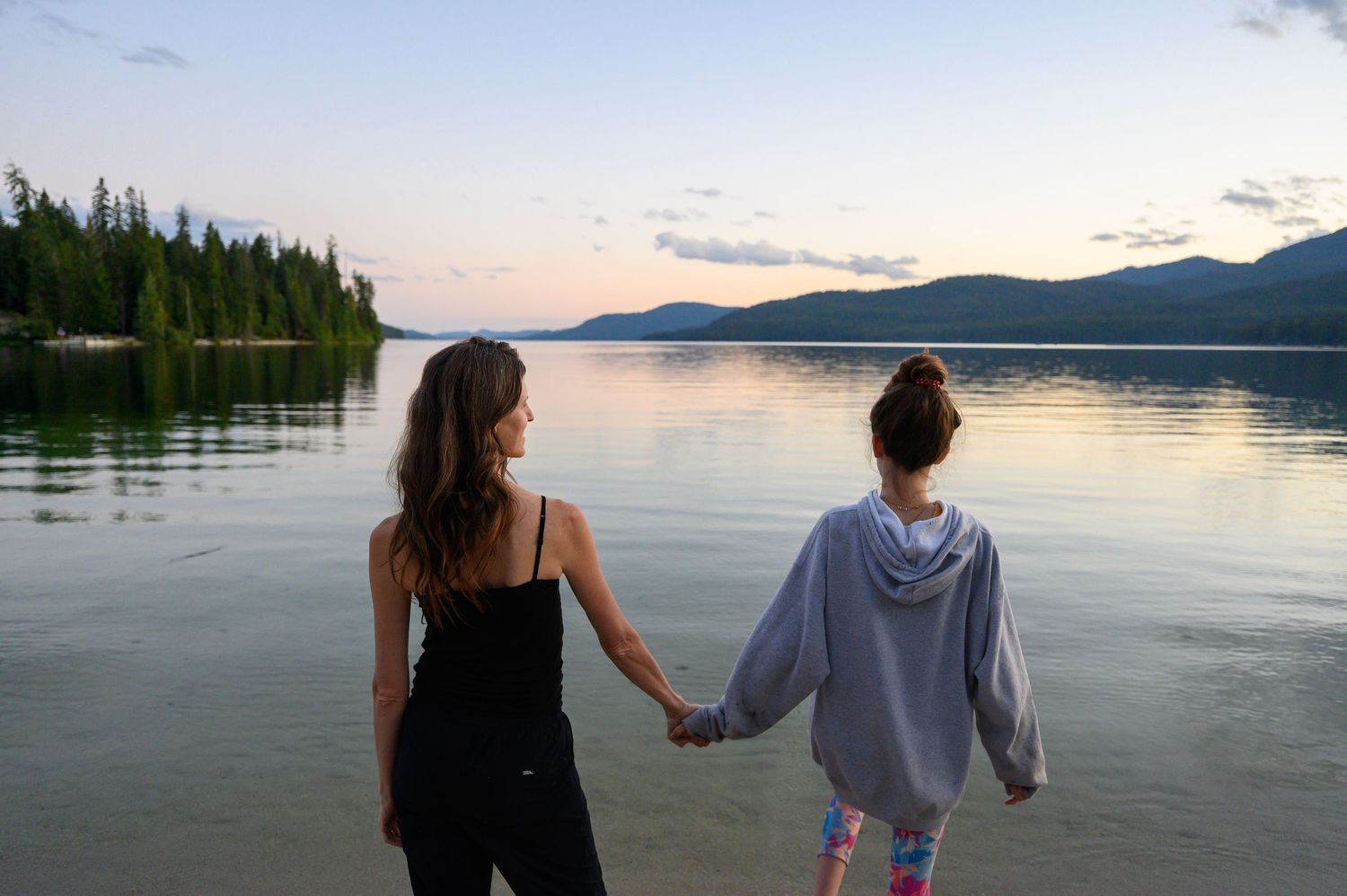 Two people stand hand in hand at the edge of a tranquil lake during sunset with mountains and forest in the distance.