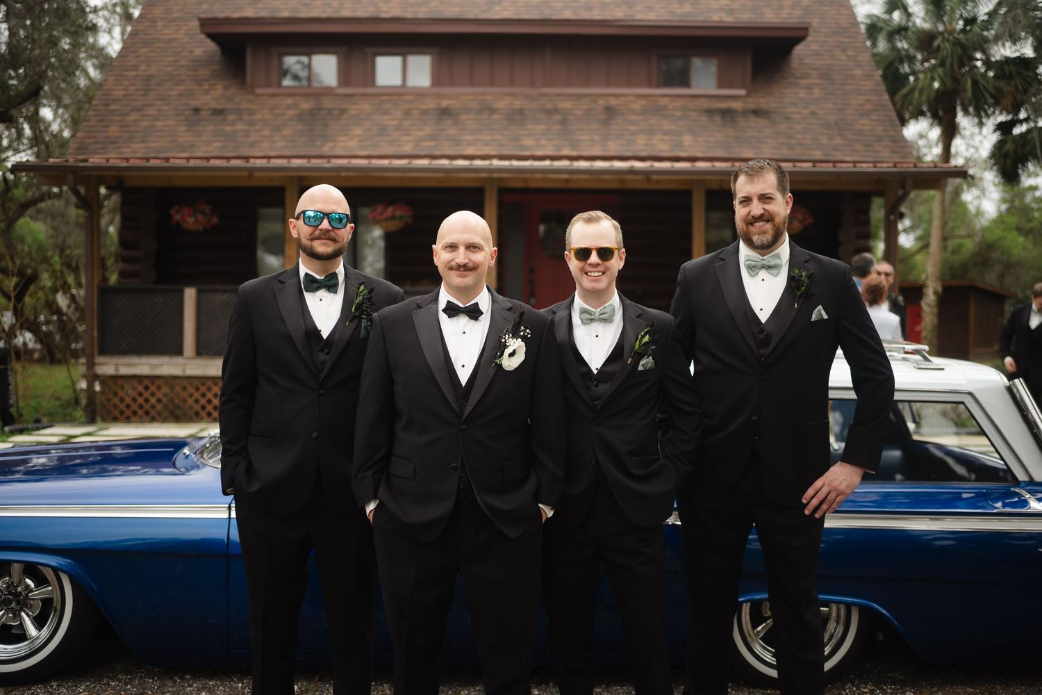 Four groomsmen in black tuxedos posing in front of a classic blue car and rustic wooden building.