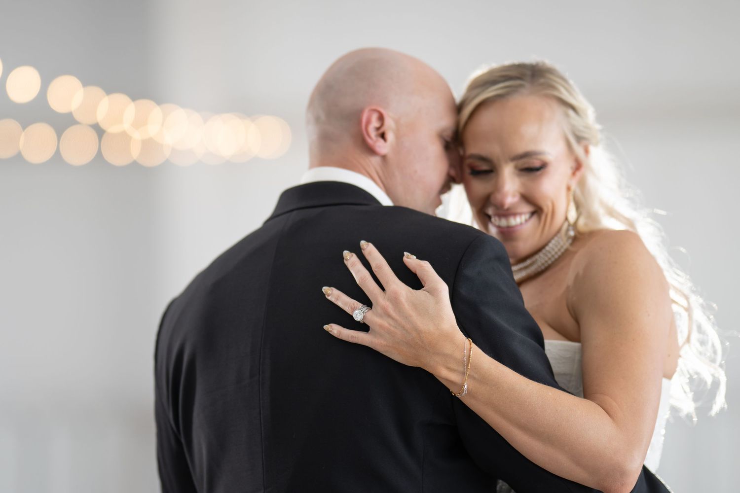 A smiling bride in a white dress embraces a bald groom in a black suit during their first dance at a wedding reception.