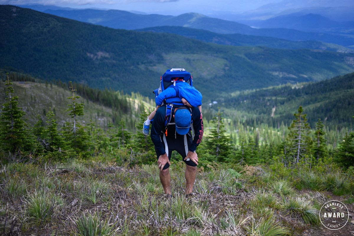 Hiker with large blue backpack trekking up a mountain trail surrounded by pine trees and scenic valley views.