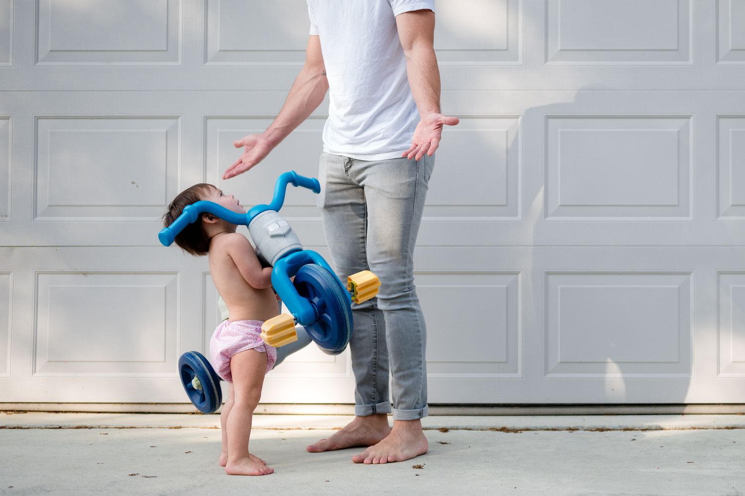 Toddler in diaper riding blue tricycle near parent standing by white garage door outdoors.