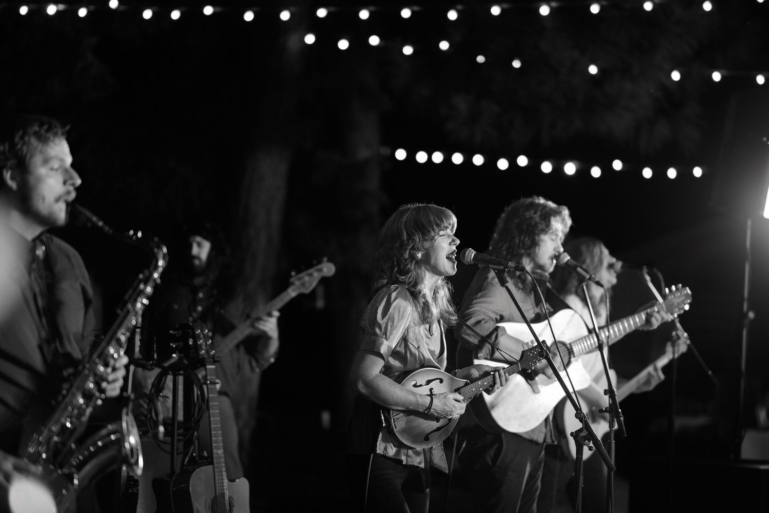 Black and white photo of musicians performing on stage with string lights overhead at a nighttime event.