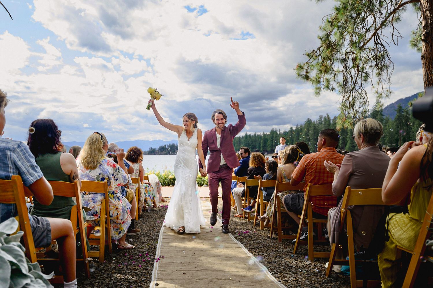Bride and groom celebrate after outdoor lakeside wedding ceremony, walking down aisle with guests seated on yellow chairs.