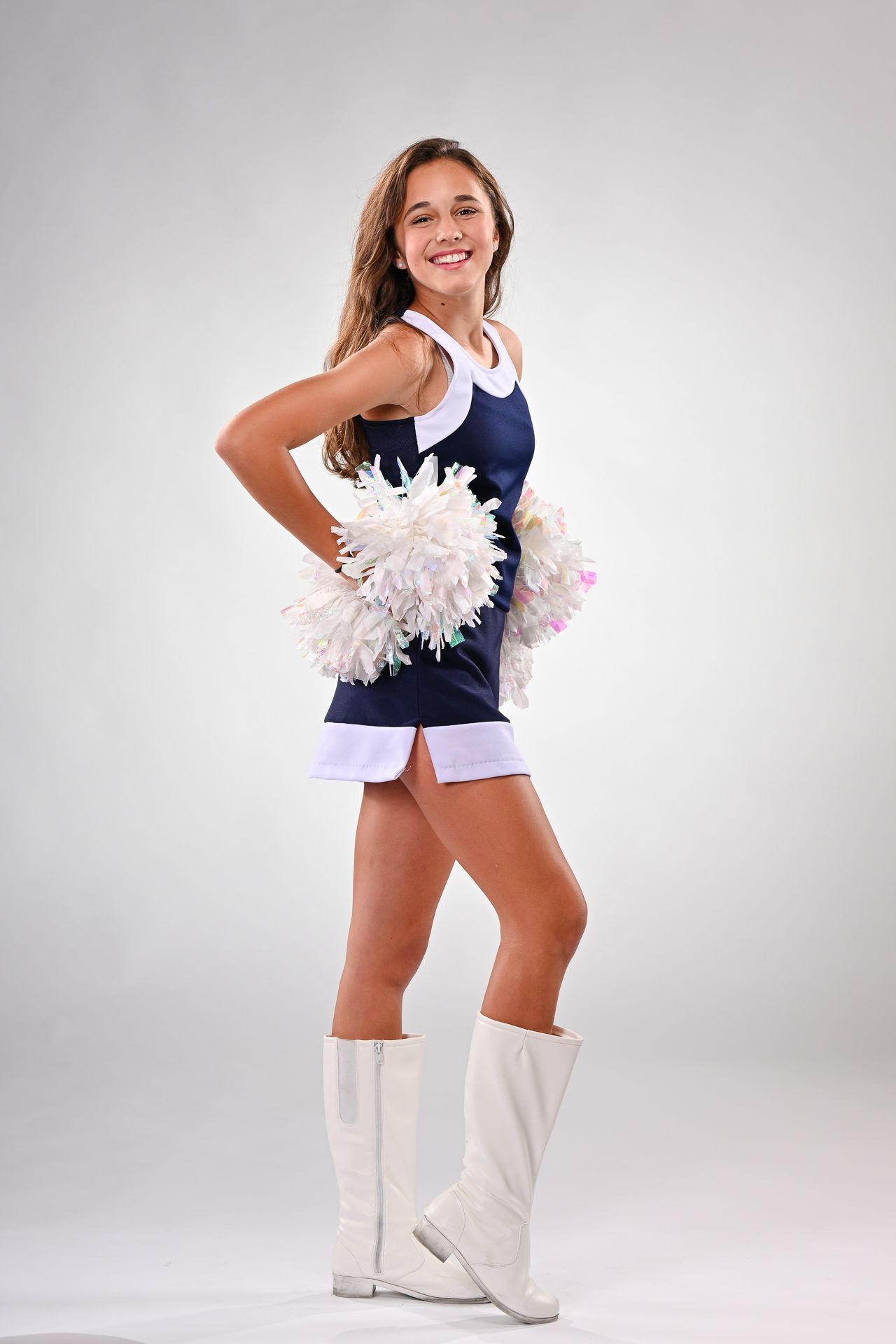 Young cheerleader in navy uniform with white pom-poms and boots poses confidently against gray background.