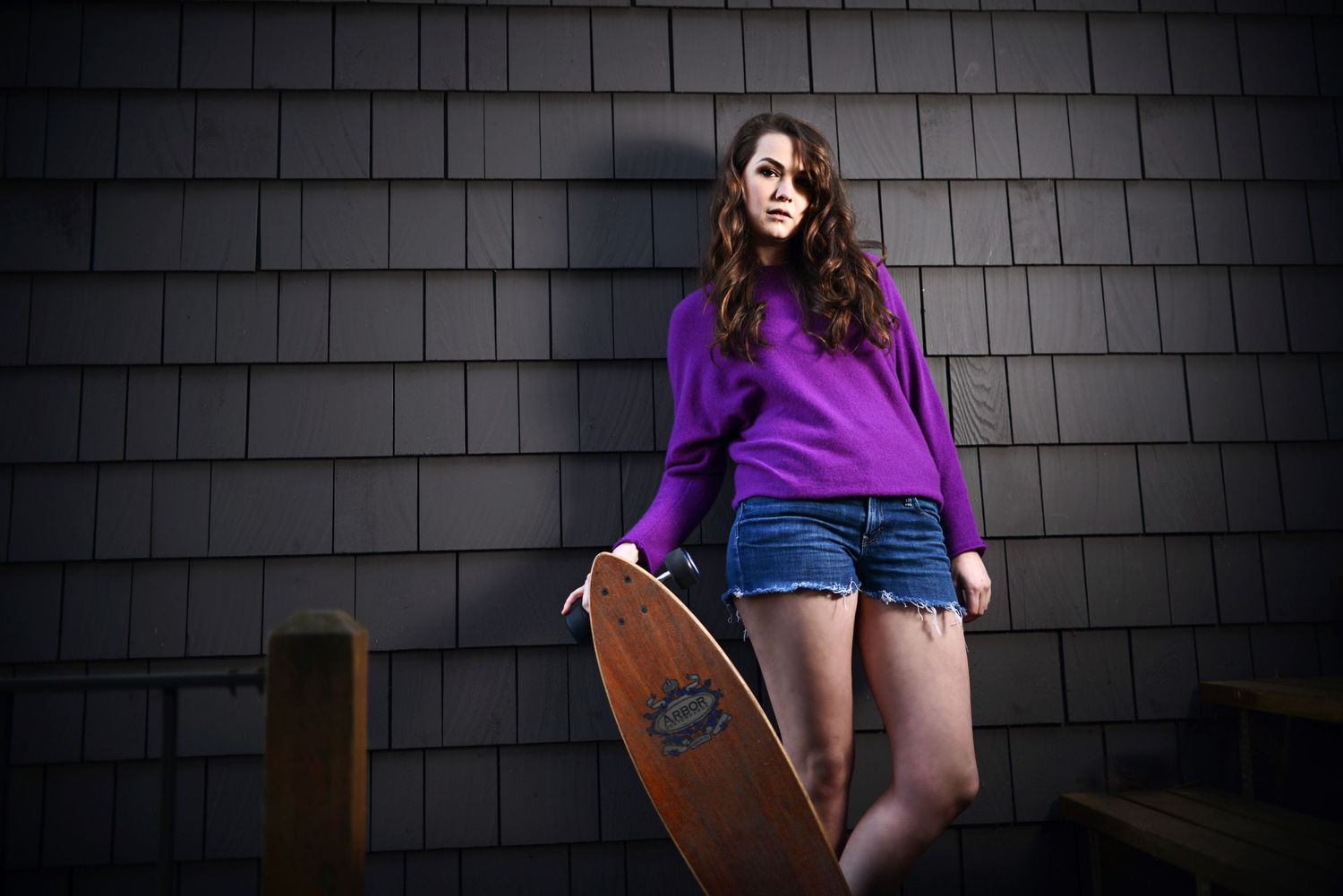 Young woman in purple sweater and denim shorts holding a wooden skateboard against a gray shingle wall.