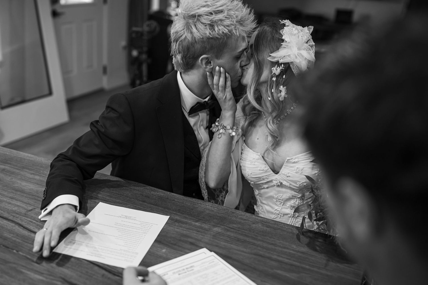 Black and white photo of a bride and groom sharing a kiss while signing their wedding certificate at a wooden table.
