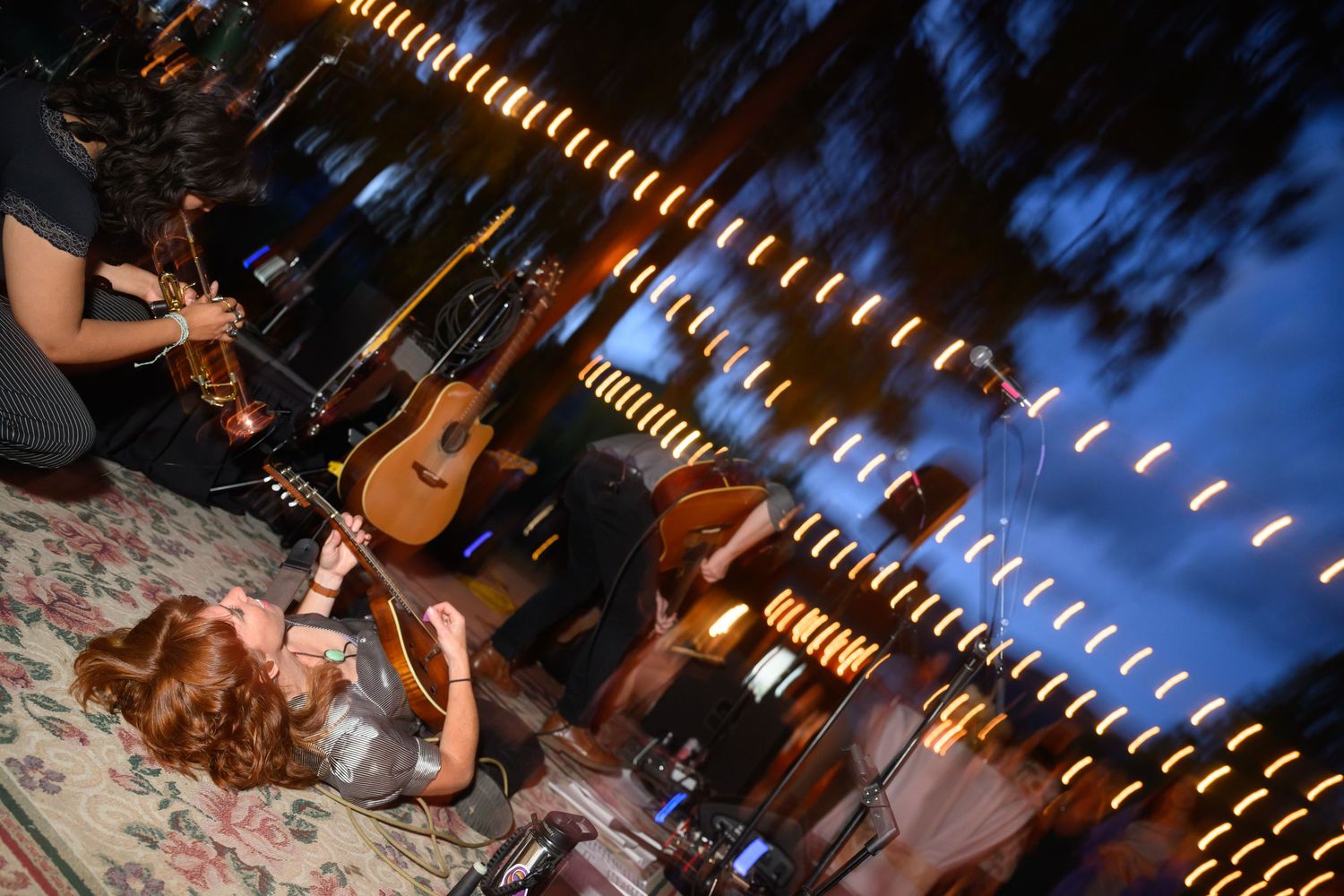 String lights create illuminated trails above a nighttime outdoor party setting with a guitar visible.