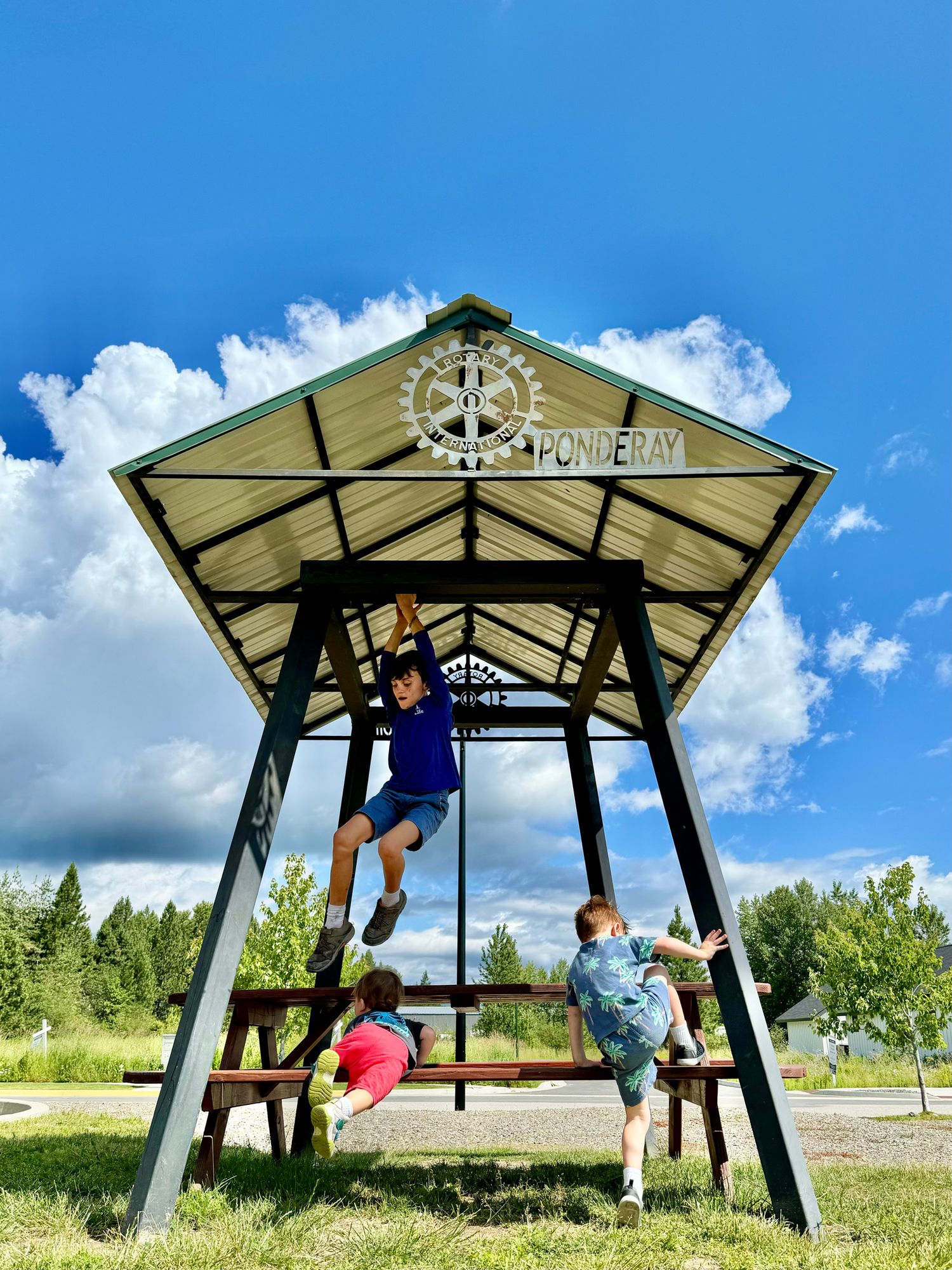 Children playing on an outdoor gazebo structure with climbing equipment under blue summer skies.