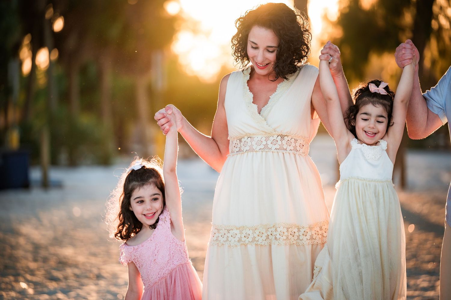 A family walks hand in hand along a beach at sunset wearing flowing dresses in soft pink and white tones.