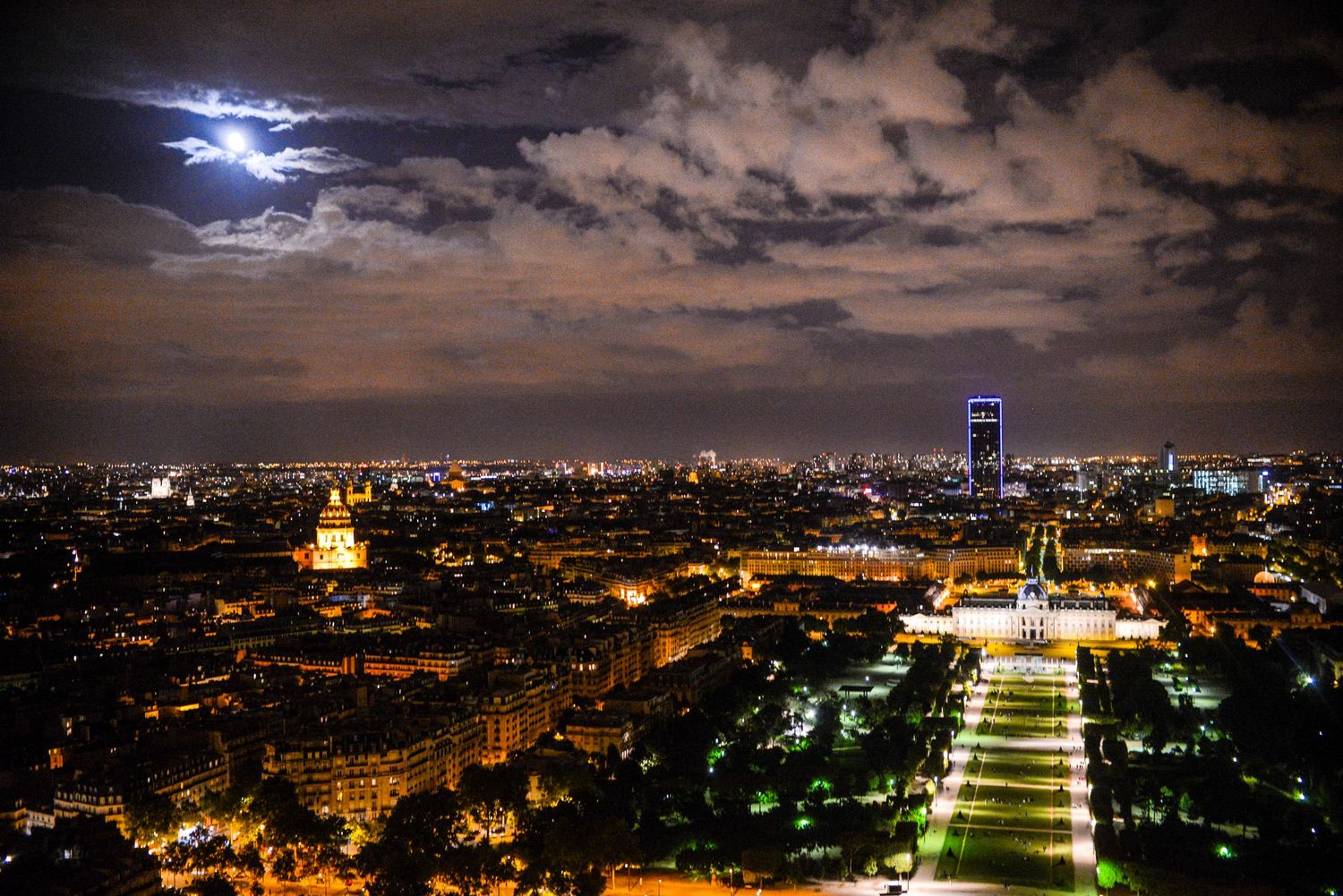 A panoramic night view of Tbilisi, Georgia shows illuminated buildings, landmarks and city lights under a cloudy moonlit sky.