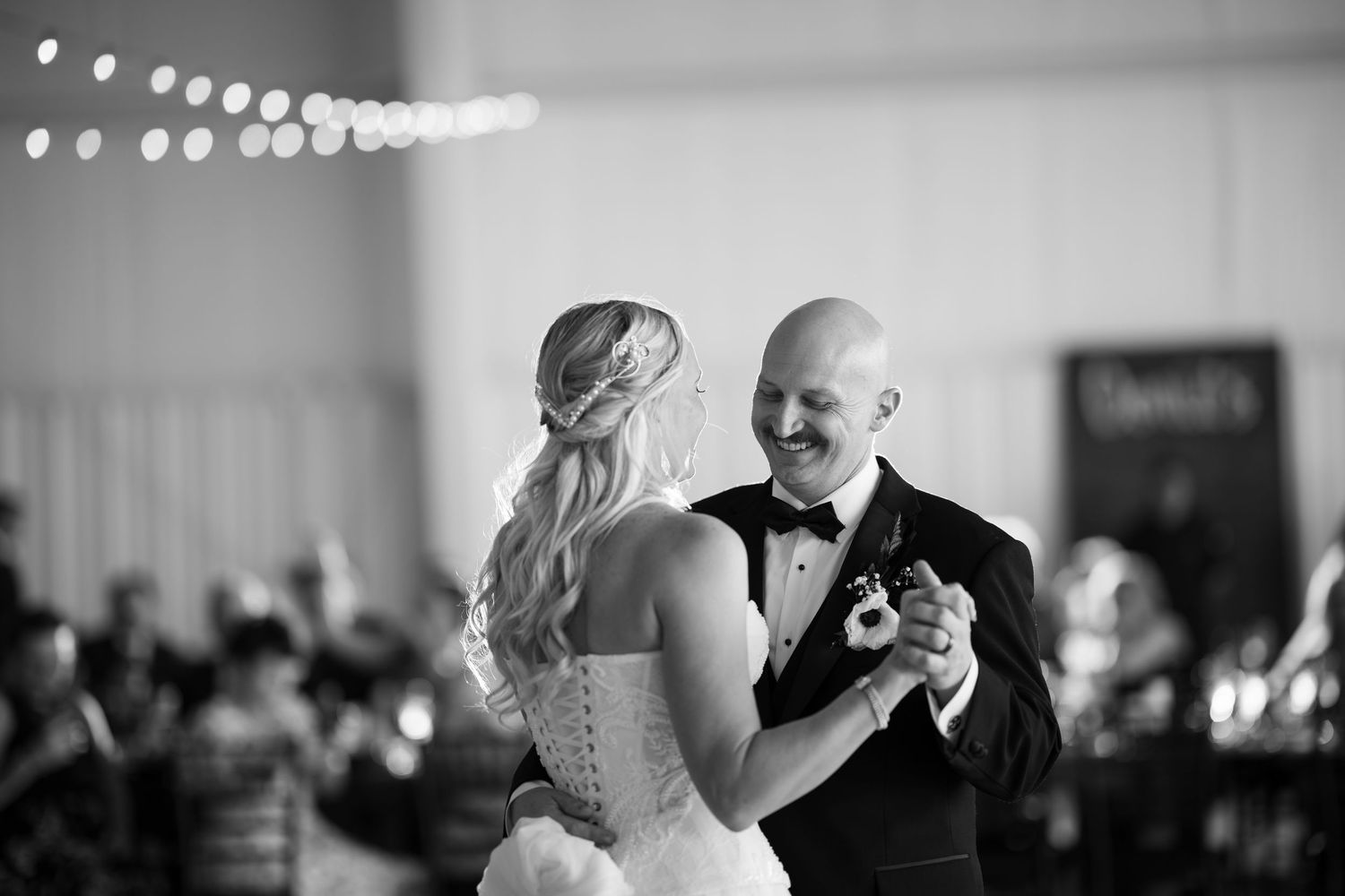Bride and groom share their first dance in black and white wedding photo with guests in background.