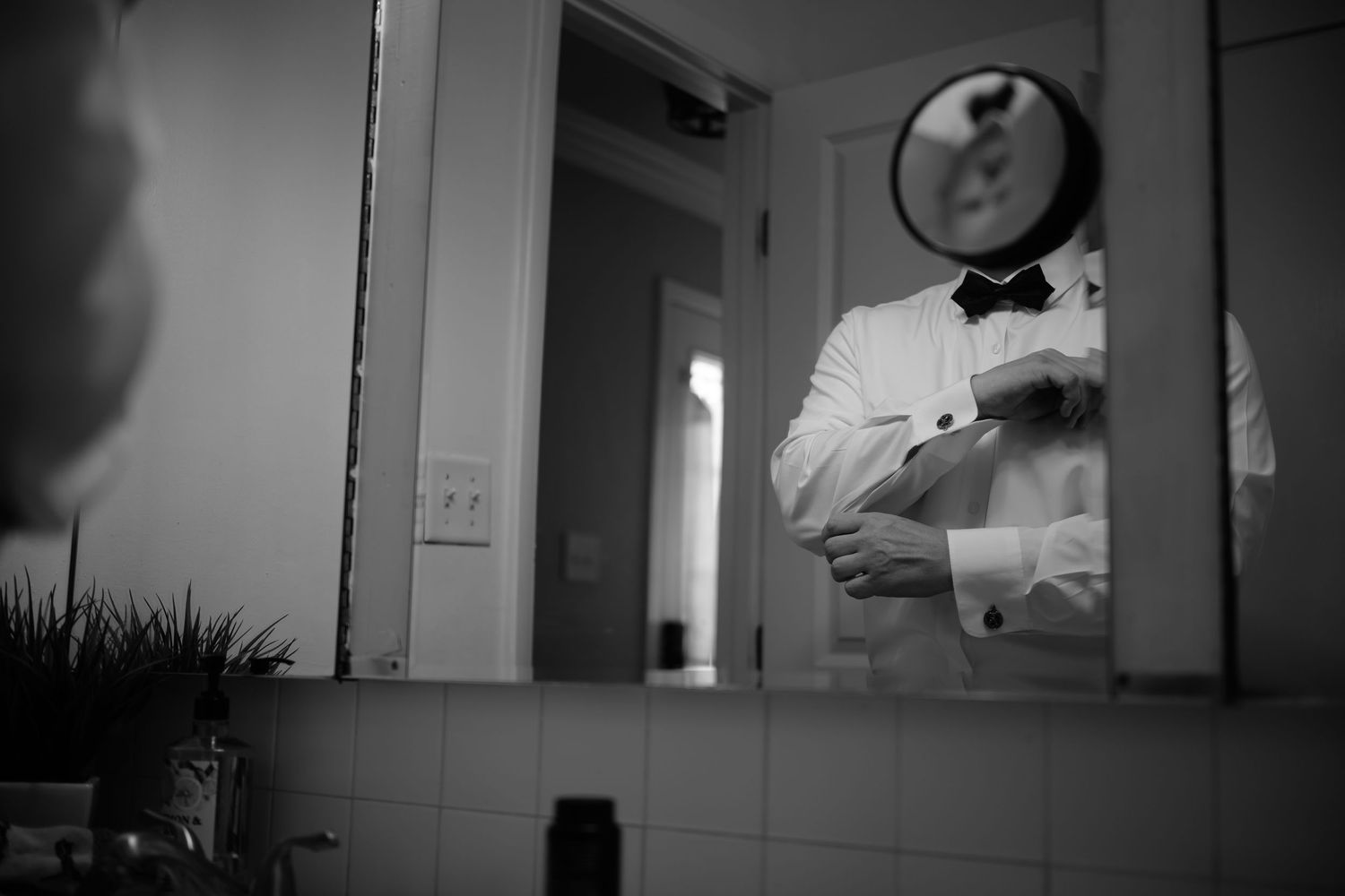 Groom in tuxedo adjusting cufflinks in bathroom mirror reflection before wedding ceremony.