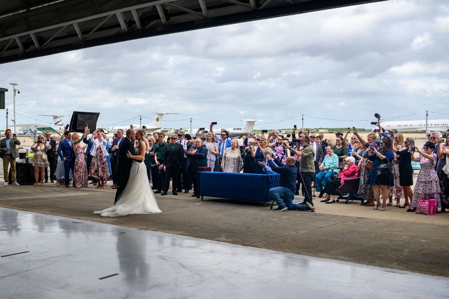 Bride and groom share first dance as wedding guests celebrate outdoors near an airport hangar.