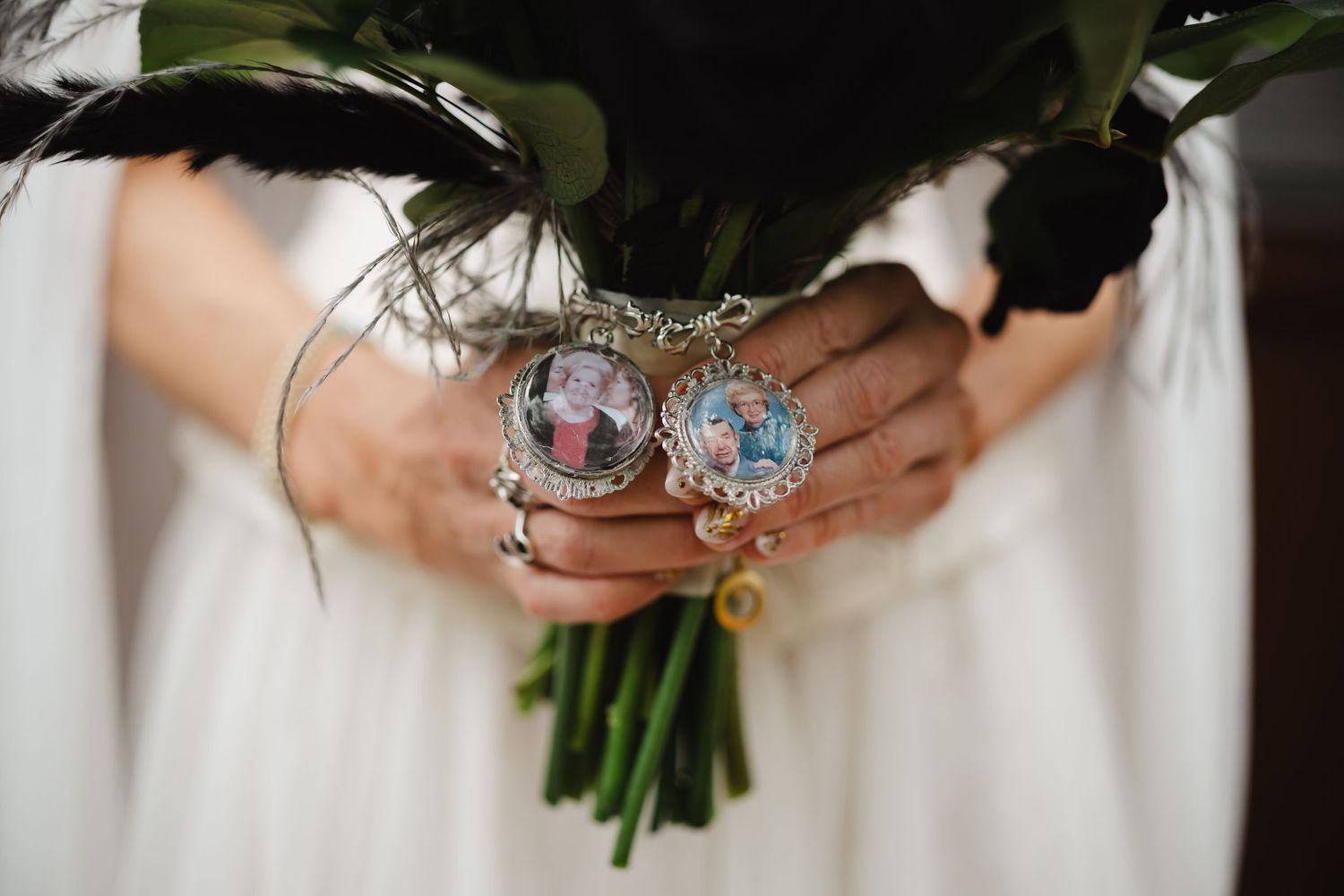 Bride holding a dark floral bouquet with memorial photo charms and rings on her fingers on her wedding day.