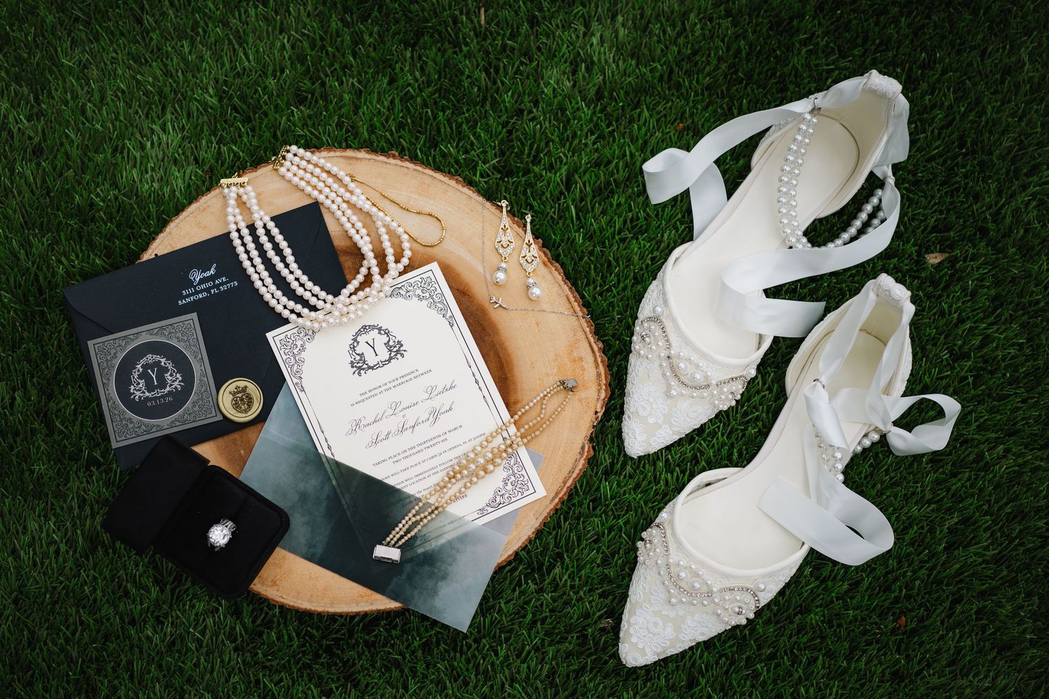 Elegant wedding flat lay with pearl necklace, diamond ring, white heels, and formal invitation on wood slice atop green grass.