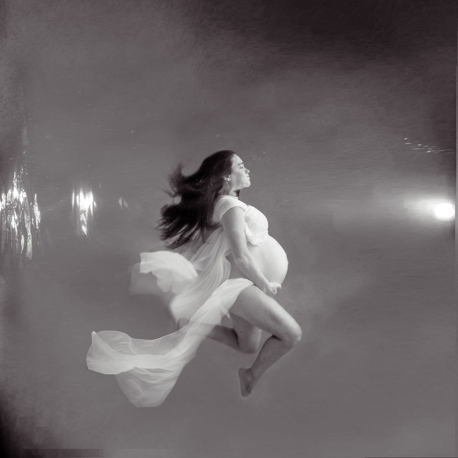 Black and white underwater photo of a pregnant woman in a flowing white dress, gracefully floating in serene water.