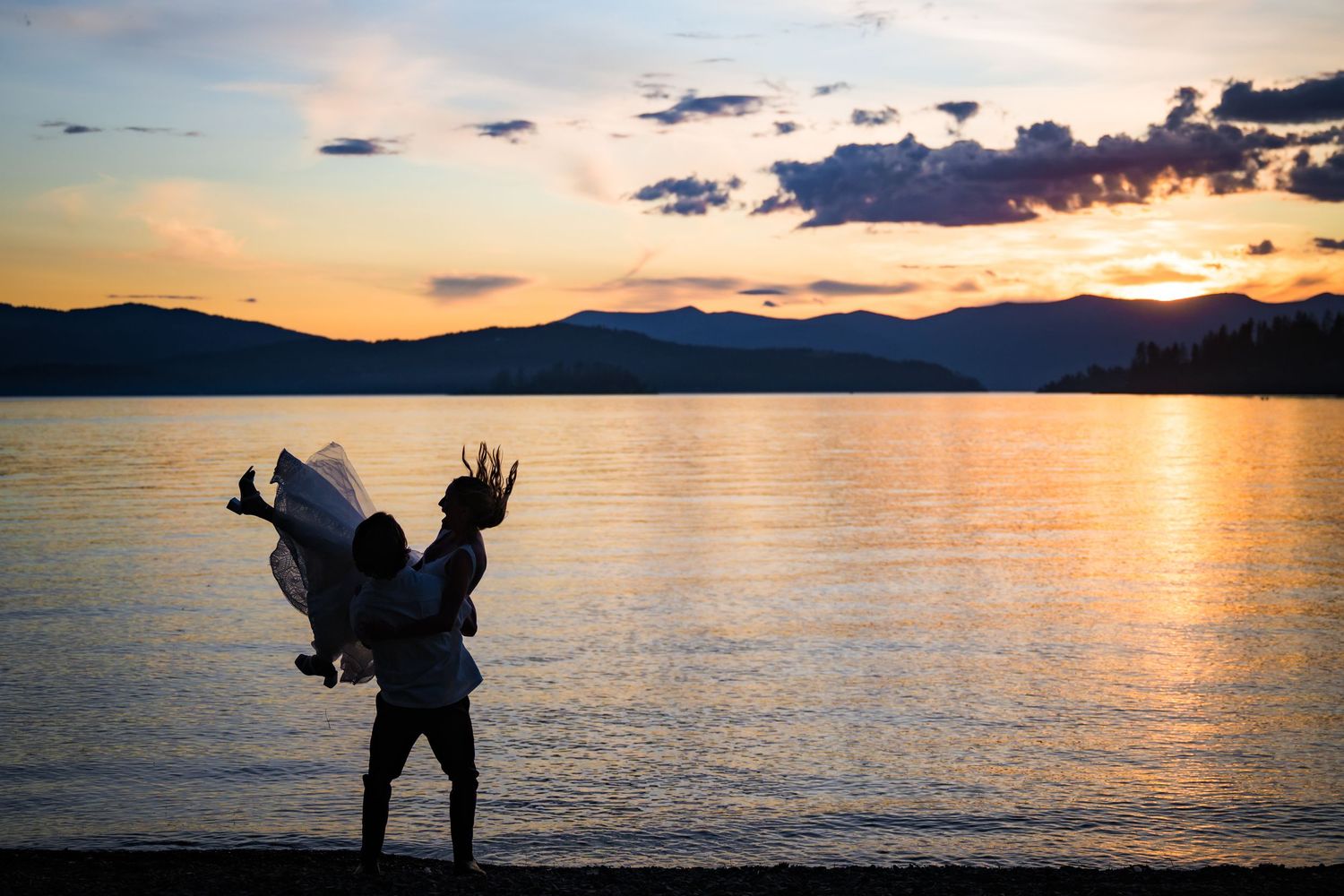 Silhouette of a person with raised arms stands at the edge of a lake during a scenic mountain sunset.