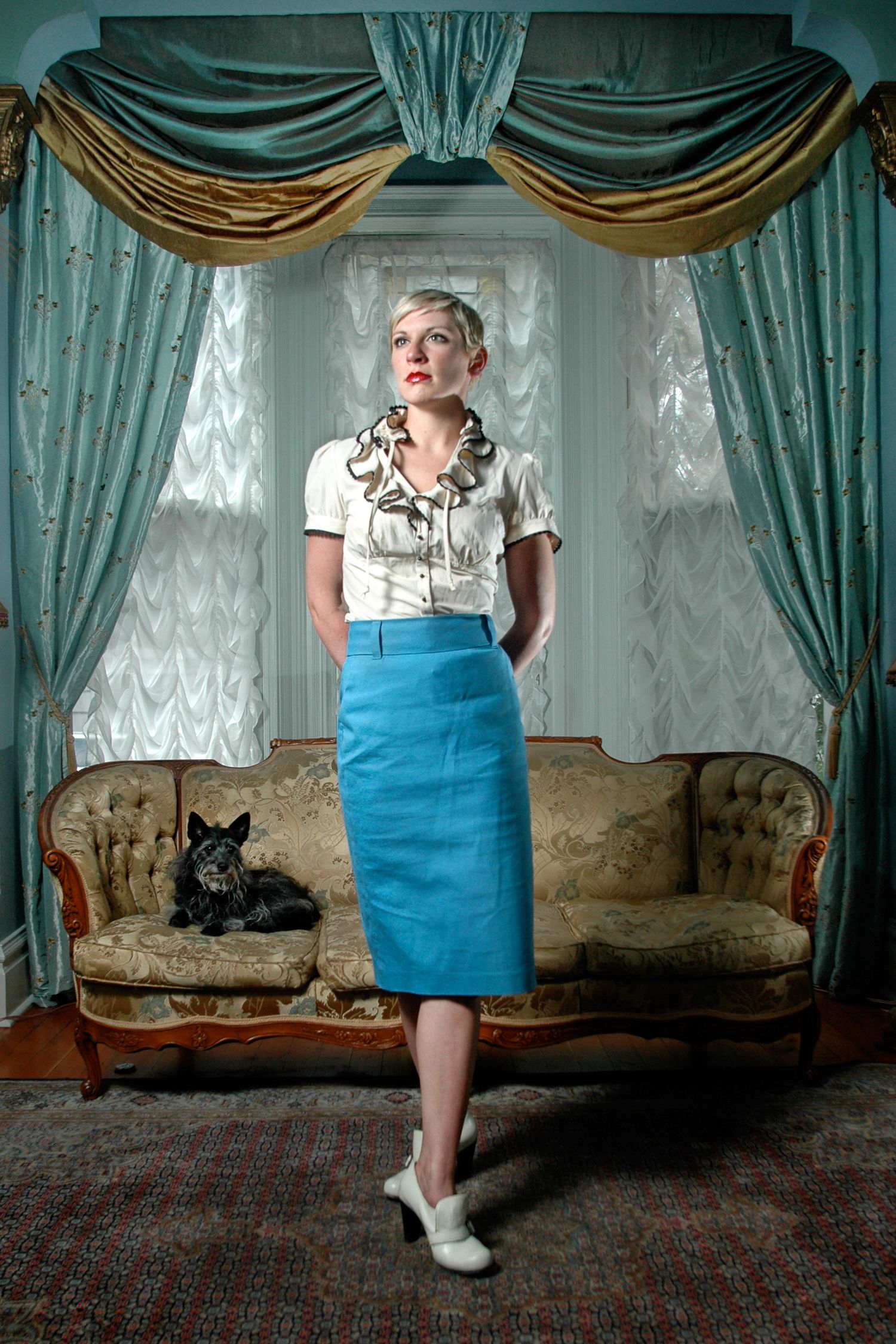 Vintage-style room with teal curtains features a person in retro outfit standing by antique couch with black cat.