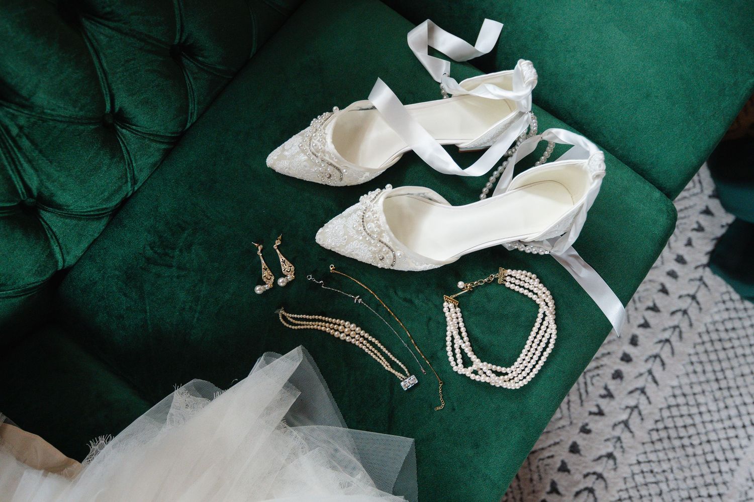 White bridal heels with pearl jewelry, earrings, and necklace displayed on a green velvet sofa.