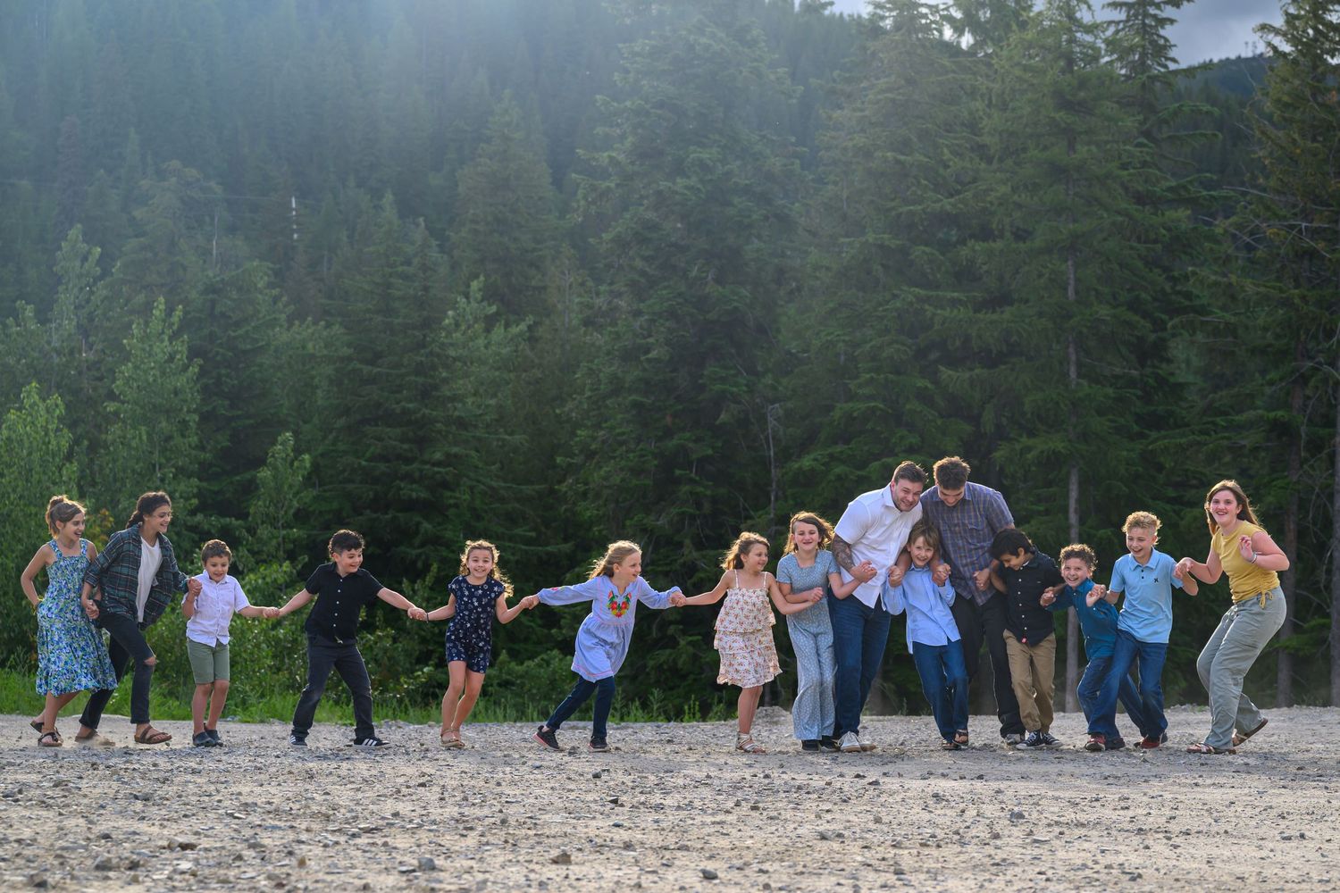 Large family holding hands in a line on a sandy beach surrounded by lush green forest trees.