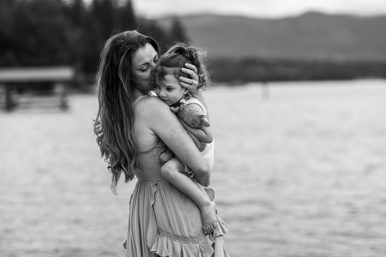 Black and white photo of a mother tenderly holding her young child near a serene lake.