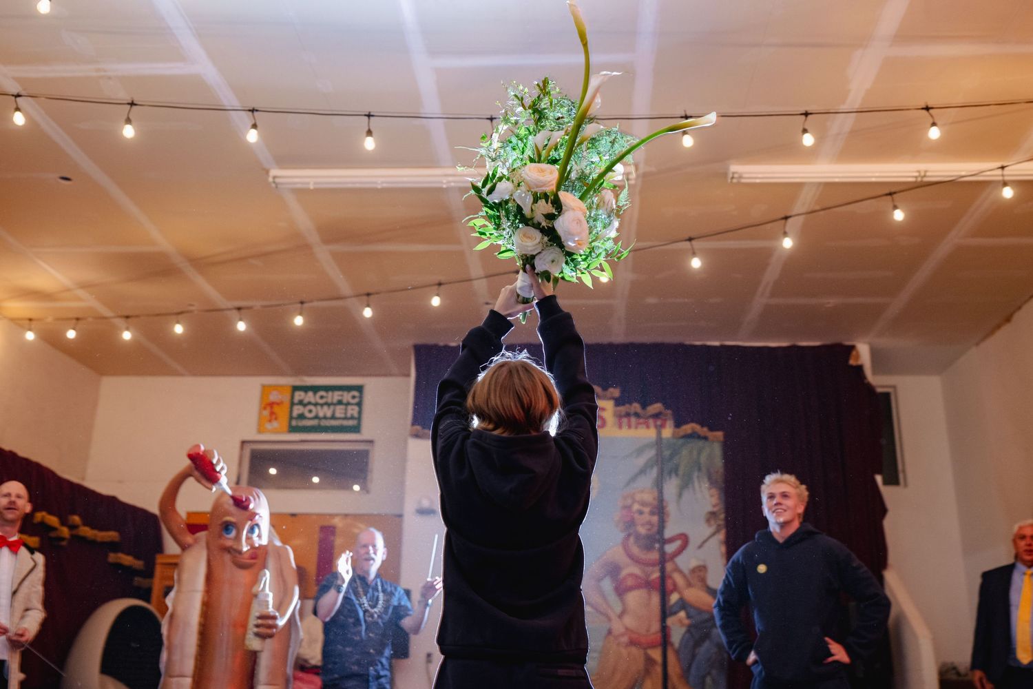 Person raising white floral bouquet overhead at indoor wedding reception with string lights.