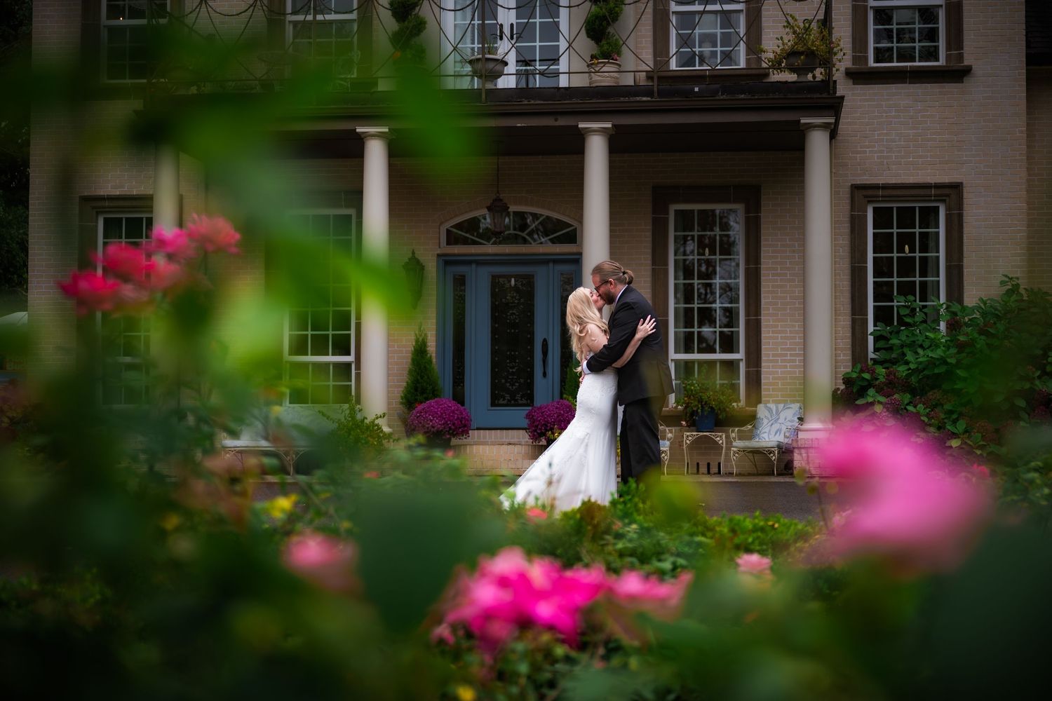 A colonial style house exterior with white trim and windows framed by pink flowering bushes and lush landscaping.