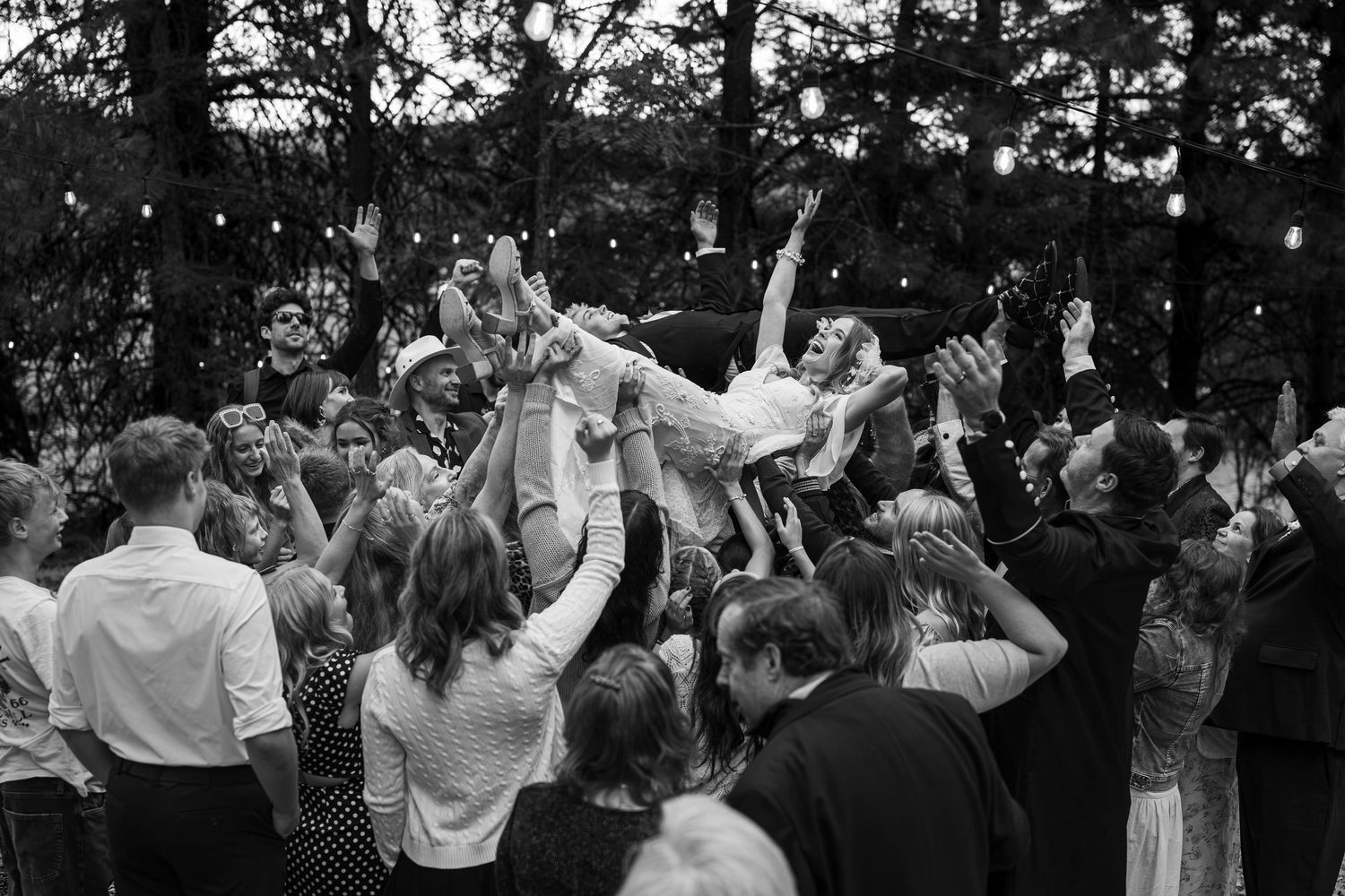 Black and white wedding crowd surf photo with guests lifting bride outdoors under string lights.