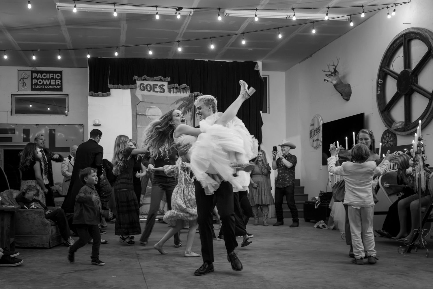 Black and white photo of couple dancing at lively indoor event with string lights and Pacific Power sign.