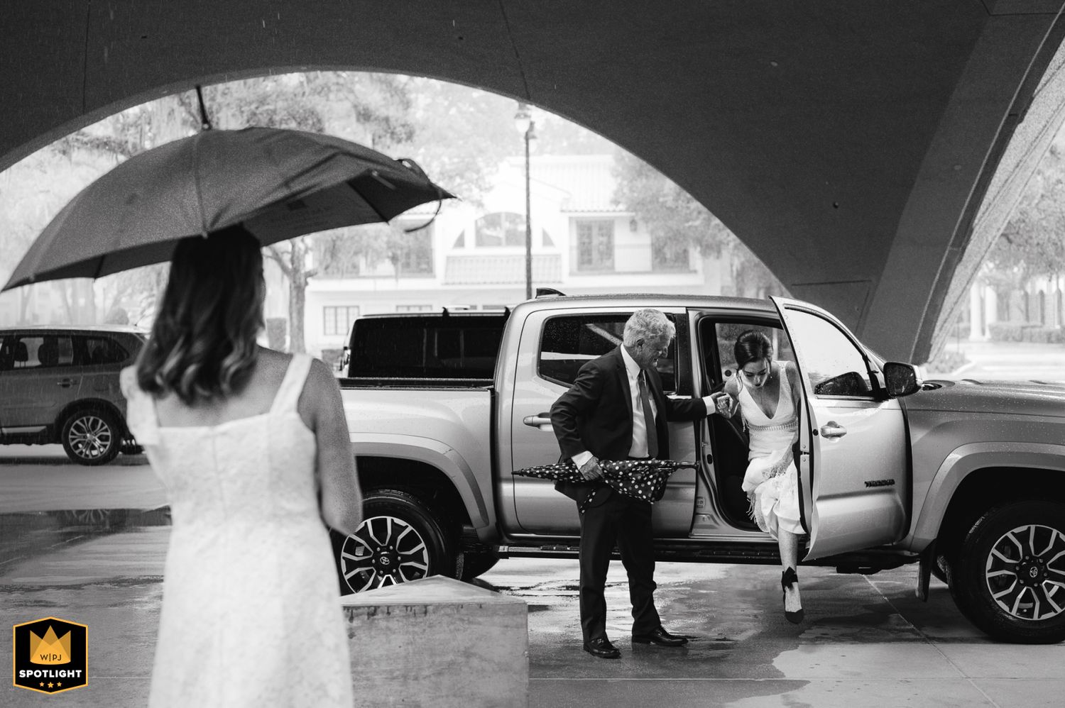 Black and white photo showing a luxury SUV parked under an archway with open doors during a rainy wedding day, father of the bride