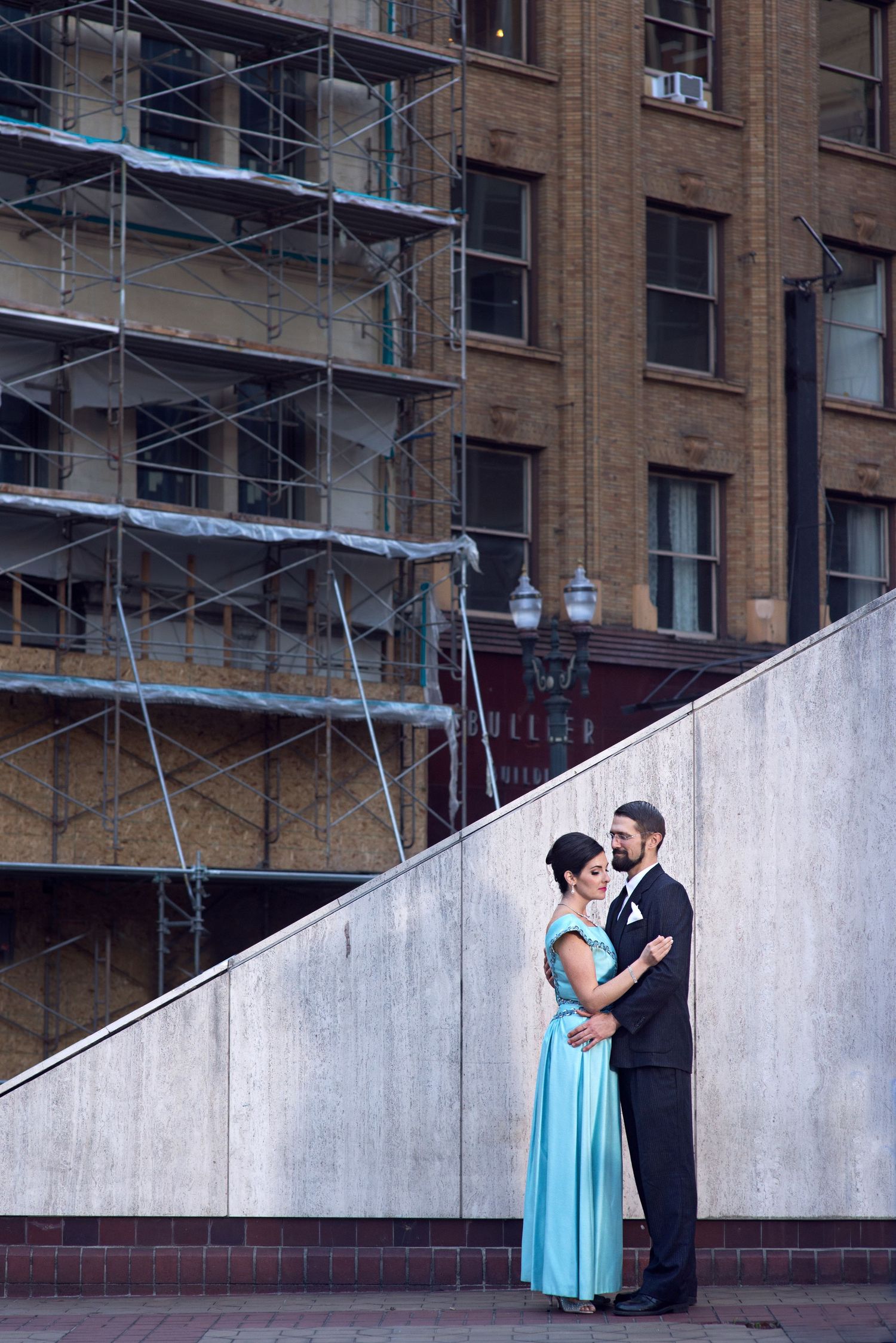 A couple in formal attire share an intimate moment against an urban brick building backdrop with scaffolding.