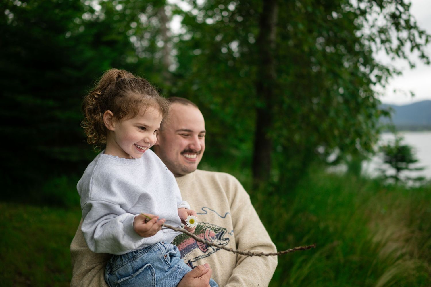 A father holds his smiling young daughter outdoors near a lake surrounded by lush green trees.
