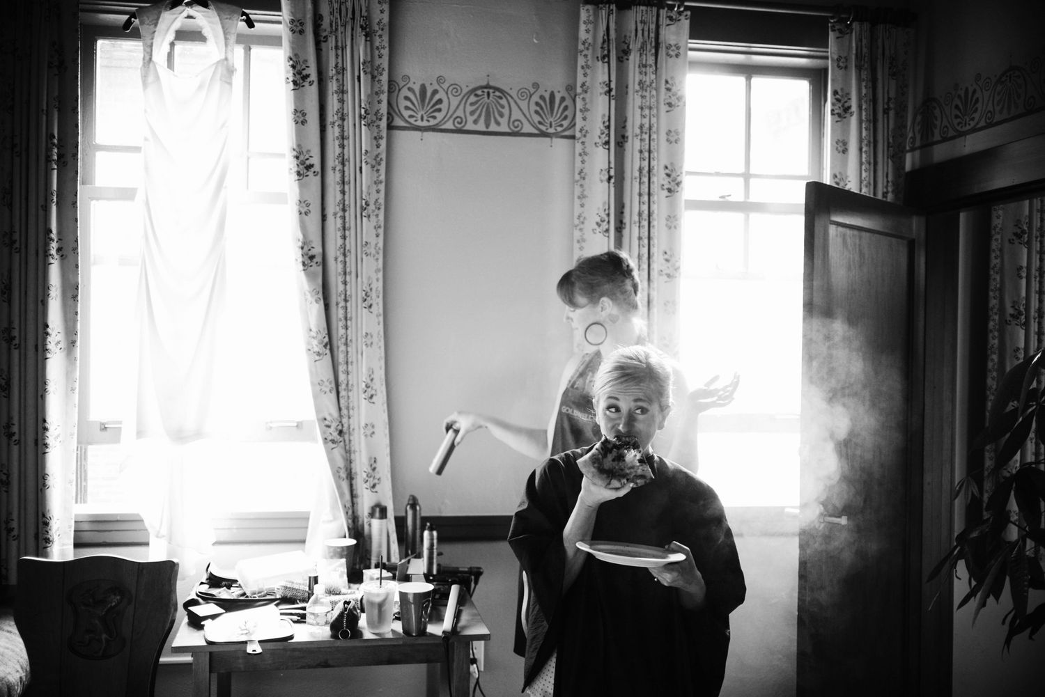 Black and white photo of two women in a sunlit room, one smoking while the other stands by a bright window.