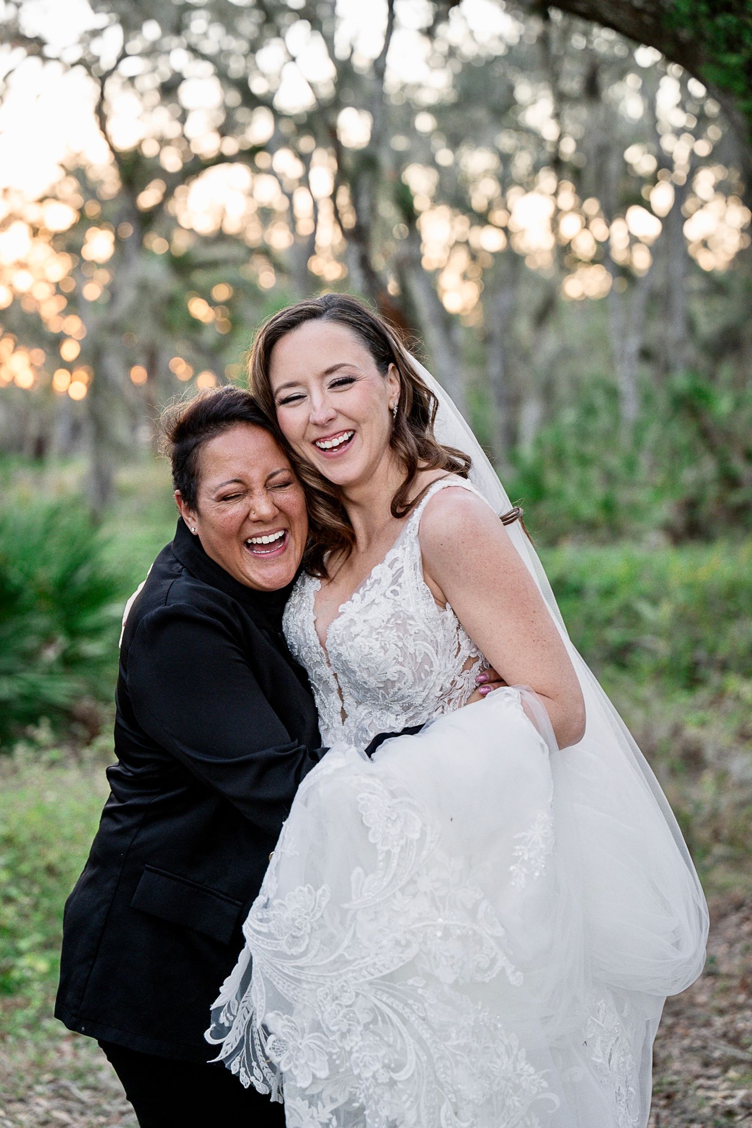 Bride and event coordinator laughing together outdoors at sunset, with one wearing an elegant lace wedding dress.