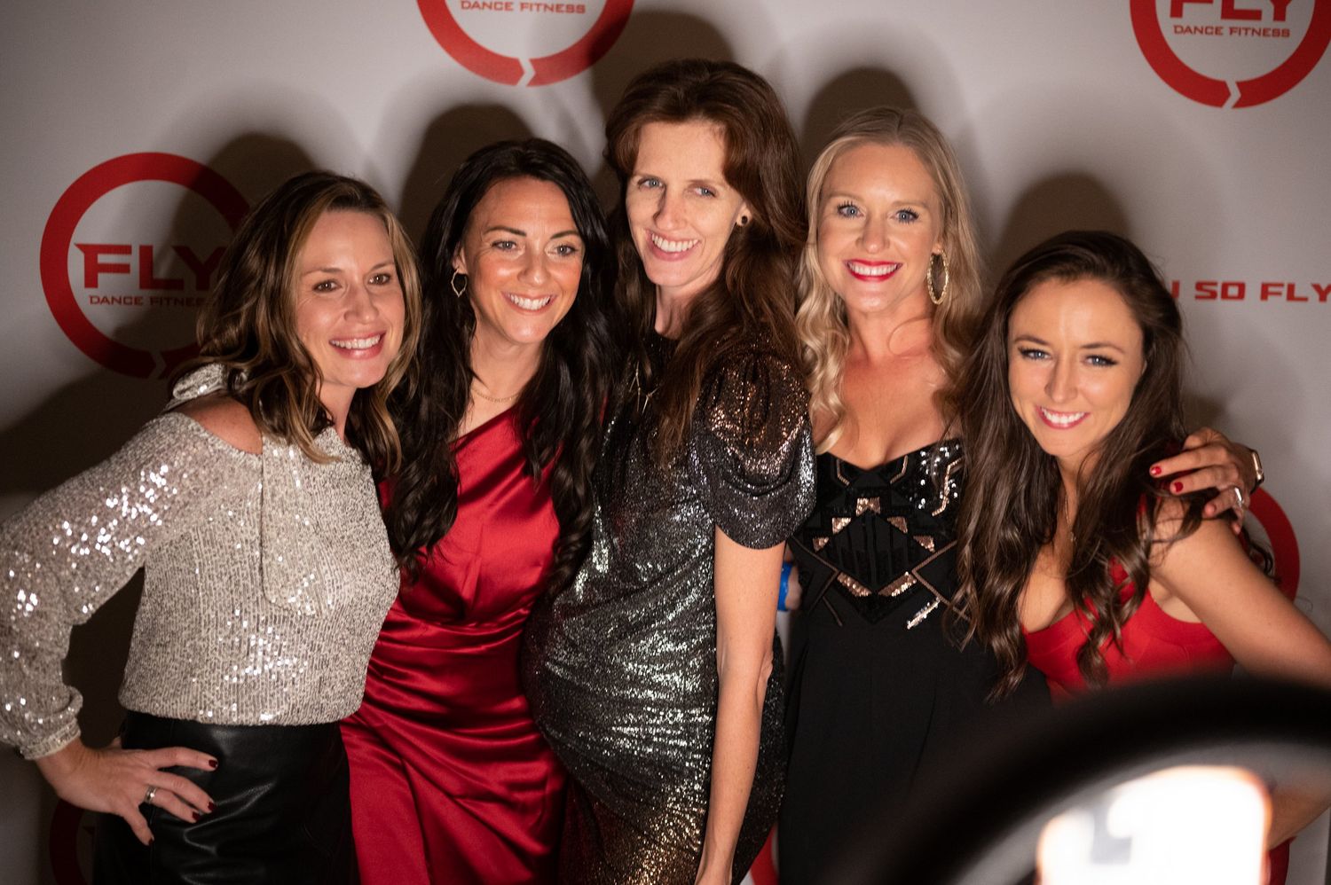 Friends posing together at an event against a branded backdrop wearing sparkly dresses.