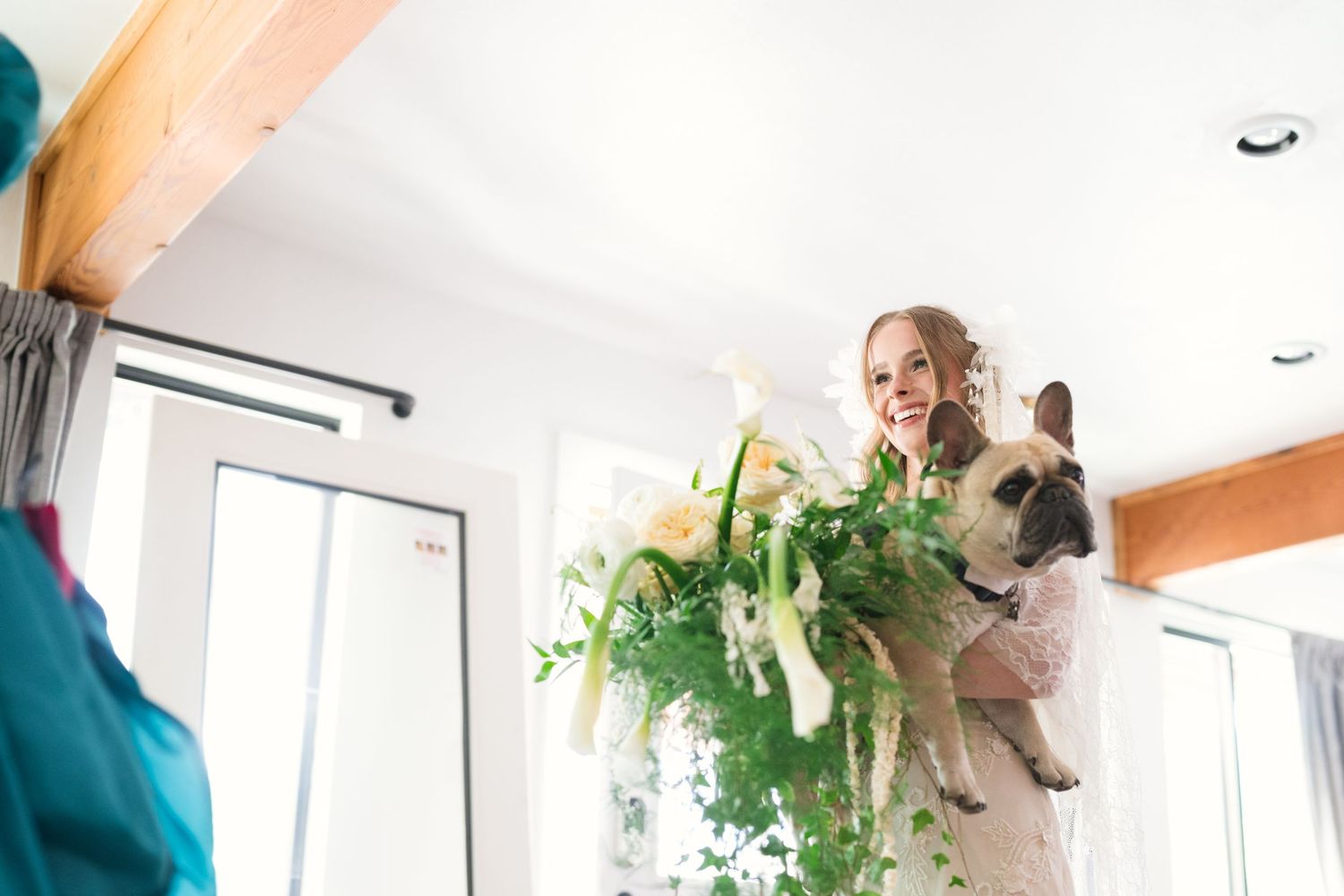 Bride in wedding dress holding a bouquet and French Bulldog, smiling in a bright white room.