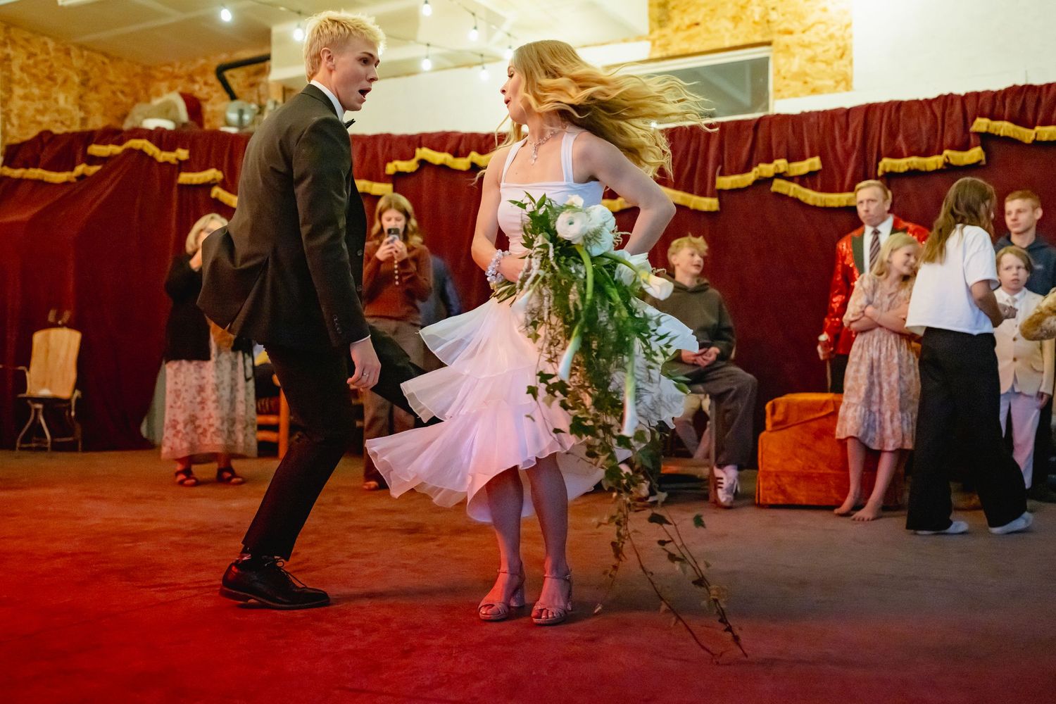 Bride and groom dancing joyfully at wedding reception, bride holding bouquet with guests watching nearby.
