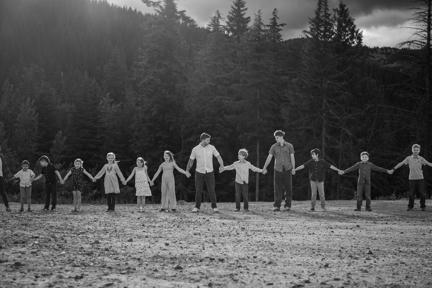 Black and white photo of children holding hands in a line outdoors with evergreen forest background.