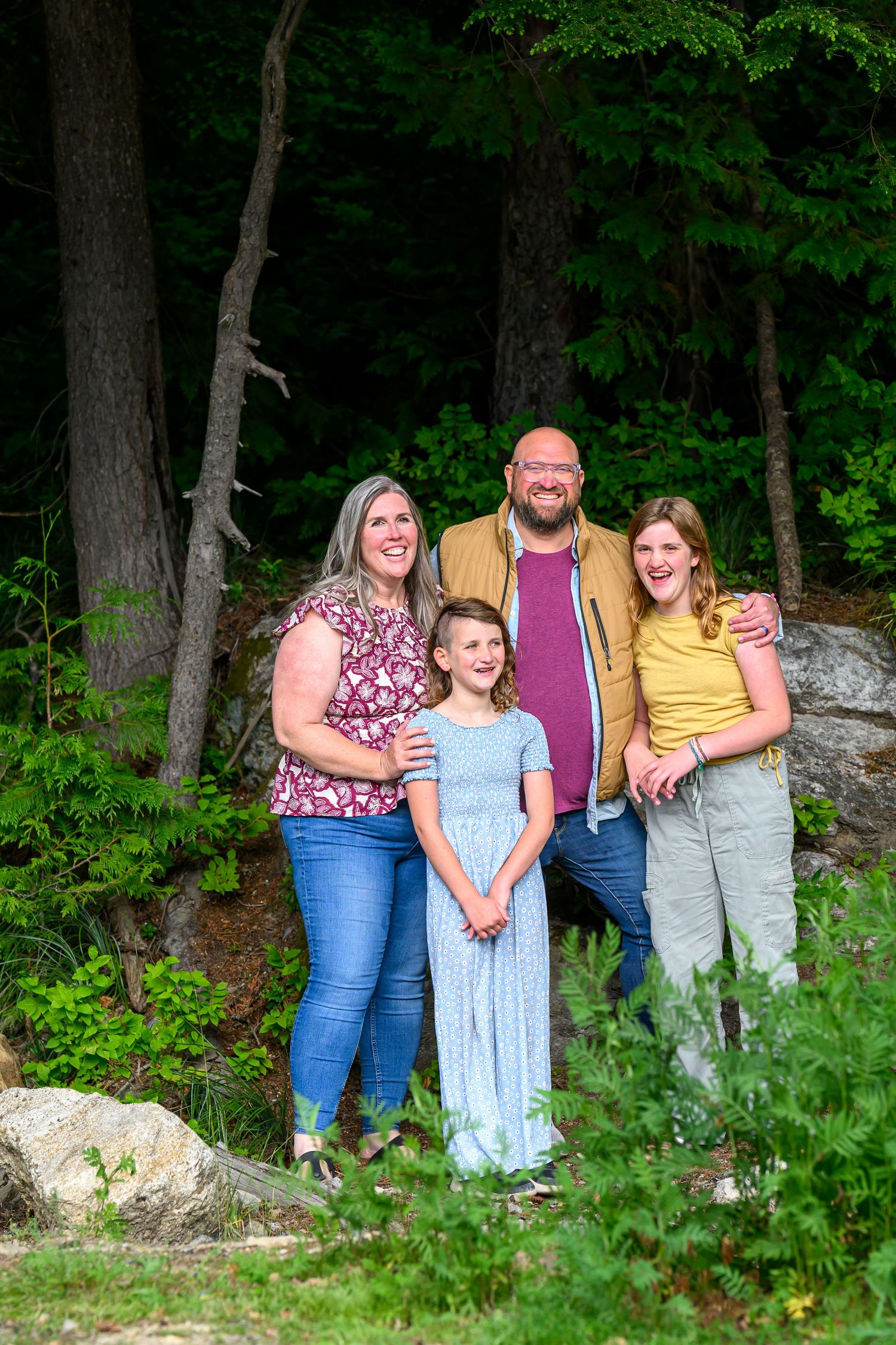 Family of four smiling outdoors in a lush green forest setting, posing together on rocky terrain.
