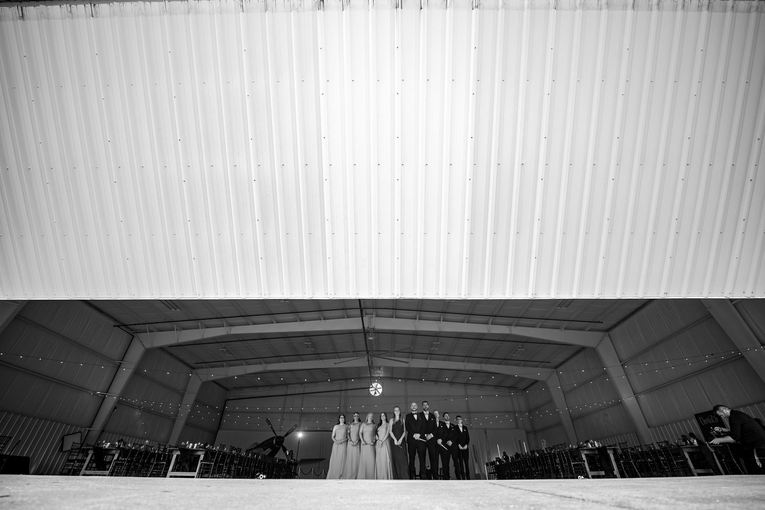 Black and white wedding ceremony photo inside a large hangar with bridal party standing at the altar.